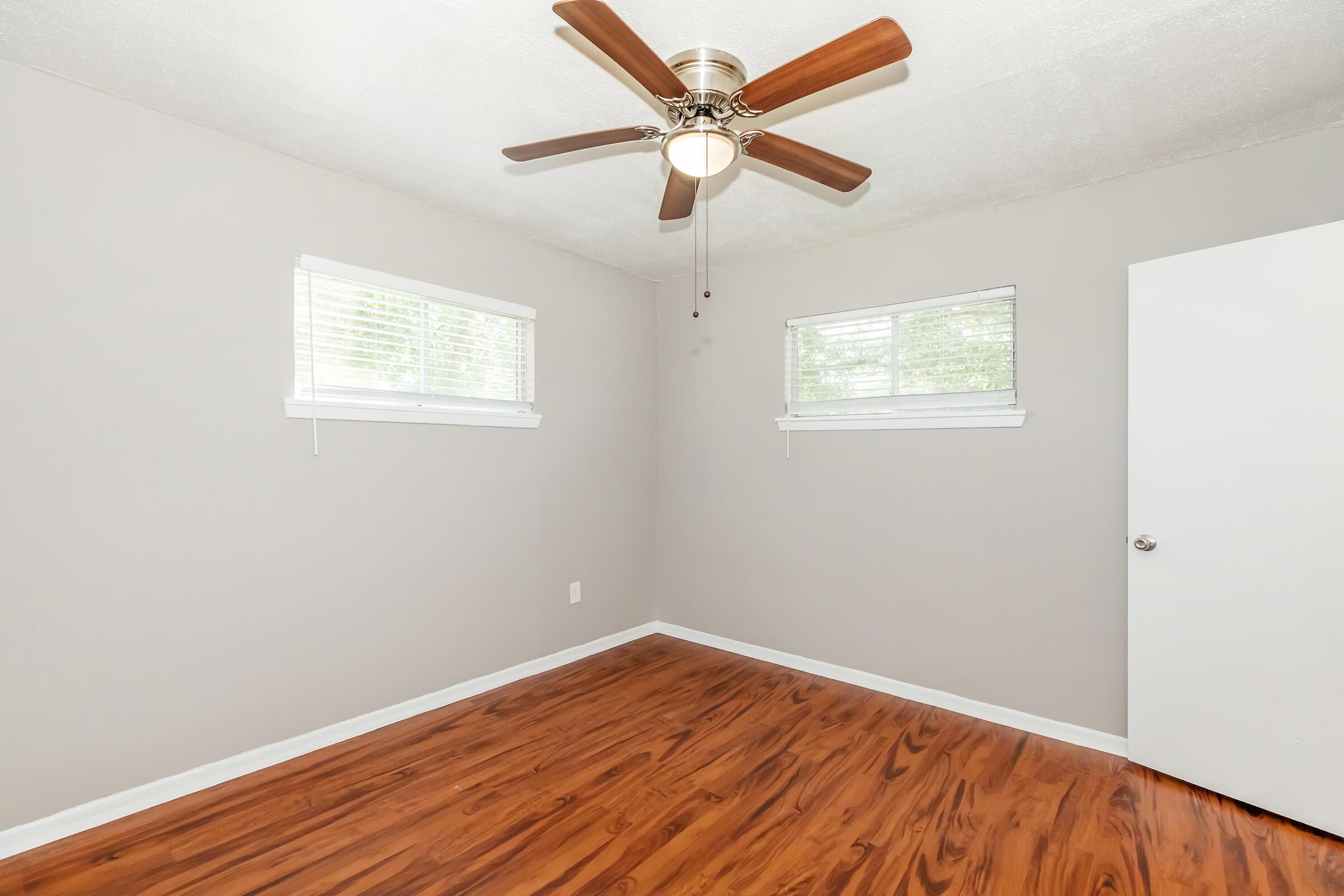 A well-lit room with light gray walls, two windows with blinds, and a ceiling fan. The floor is made of polished wooden planks, and there's a white door on the right side. The space appears empty and clean, offering a simple and airy atmosphere.
