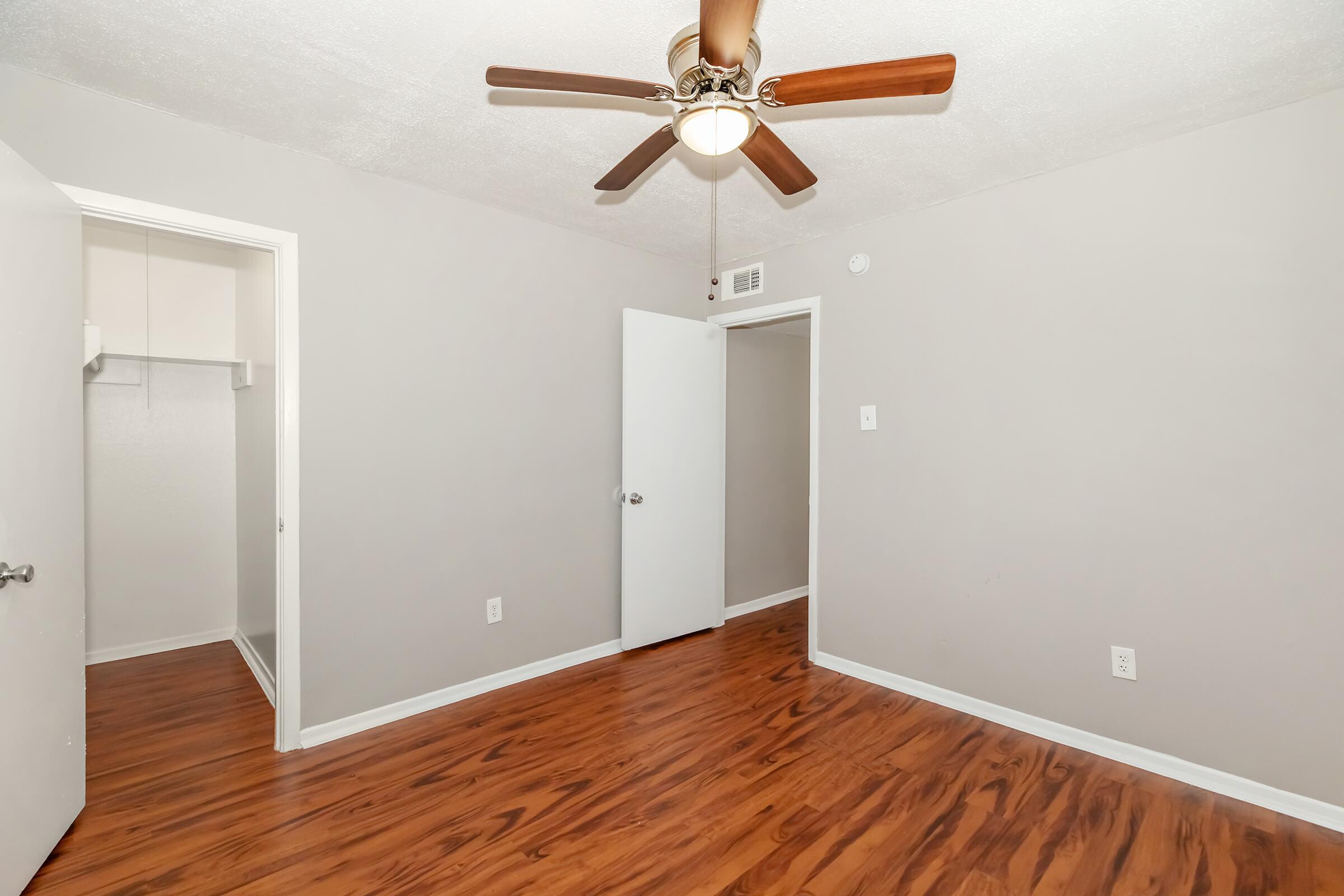 A light gray-walled room with a ceiling fan, featuring wooden flooring. There are two white doors, one leading to a closet and the other to another room. The room appears spacious and well-lit, with no furniture present.