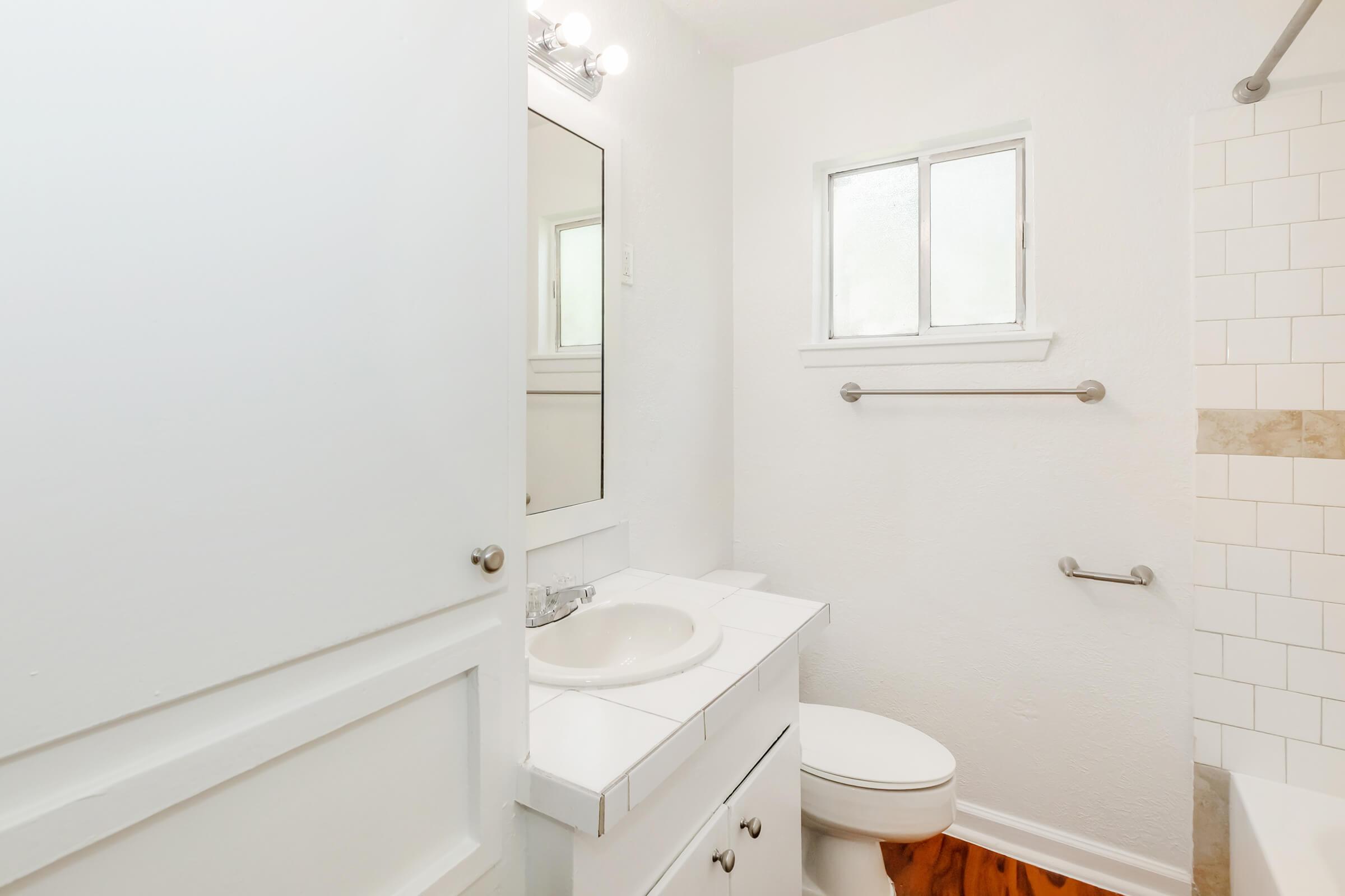 A clean, bright bathroom featuring a white sink with a large mirror above it, a toilet, and a window providing natural light. The walls are painted white, and the floor has wooden tiles. The shower area is tiled with light-colored tiles, creating a fresh and airy atmosphere.