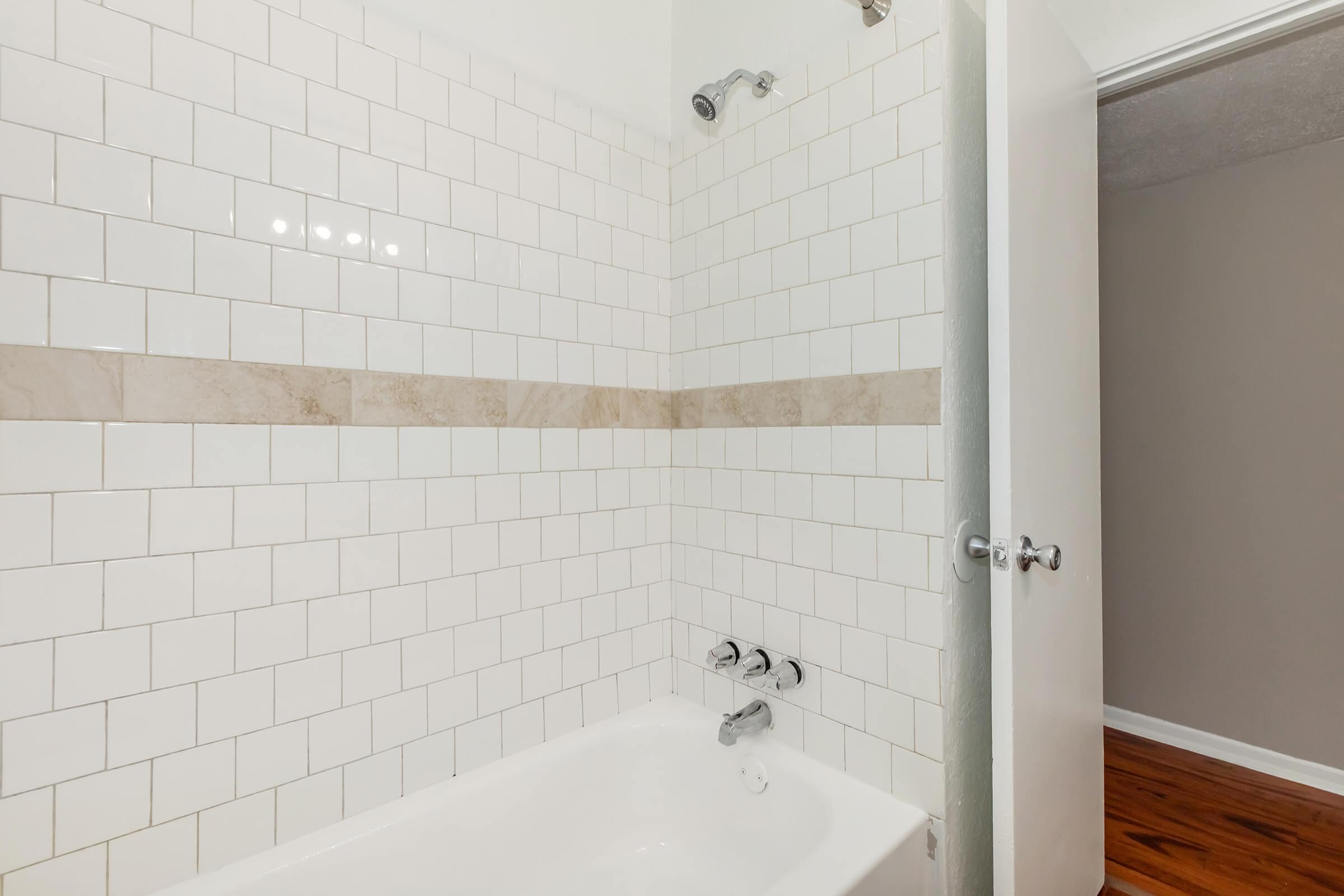 A clean, modern bathroom featuring a white tiled shower area with a light brown accent strip. The bathtub is white, and there are chrome fixtures for the shower and tub. A partially open door leads into another room with a neutral wall color and wood flooring.