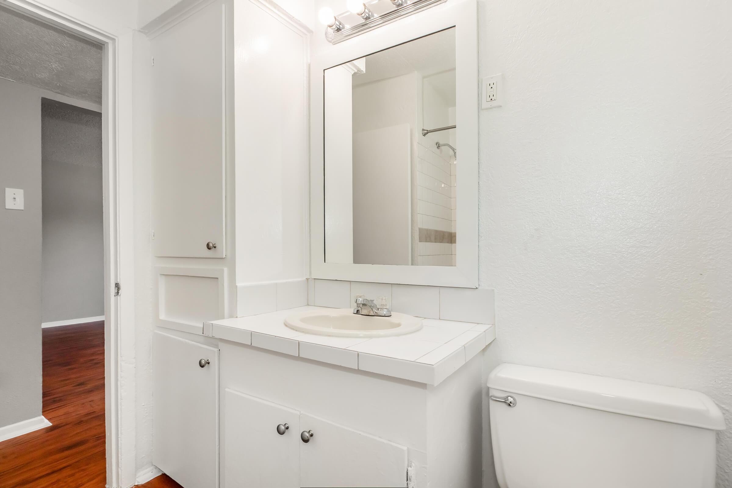 A bright bathroom featuring a white vanity with a sink and mirror above, a toilet on the right, and a light-colored wall. In the background, a glimpse of a shower area is visible, with hardwood flooring in the adjoining space.