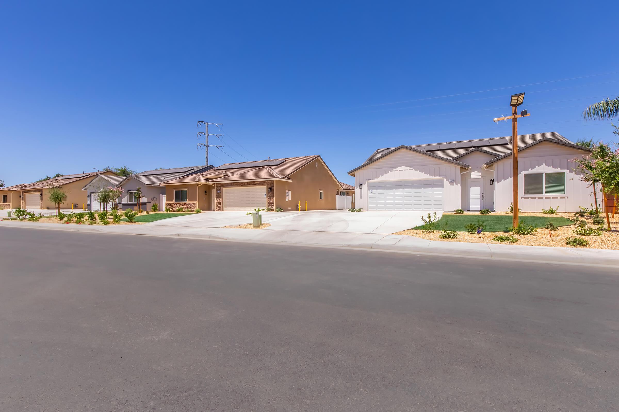 A quiet suburban street with newly constructed homes on either side. The neighborhood features well-maintained yards and driveways, under a clear blue sky. Power lines are visible in the background, adding to the peaceful residential atmosphere.