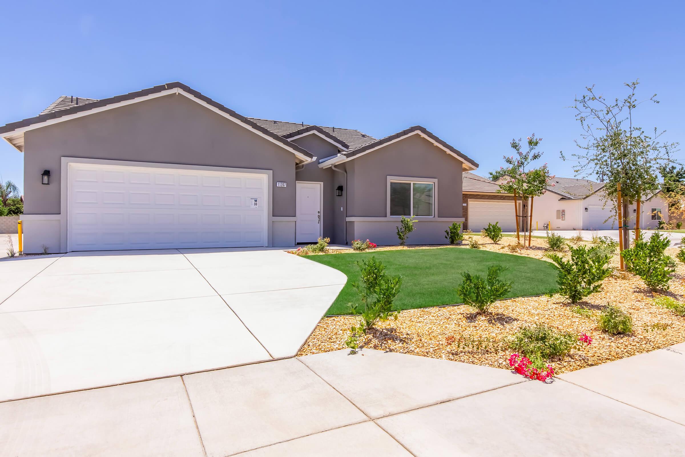 A modern house with gray exterior, a white garage door, and a neatly landscaped yard featuring green grass and small flowering plants. The driveway is wide and concrete, leading up to the entrance of the home. The background shows additional similar homes under a clear blue sky.