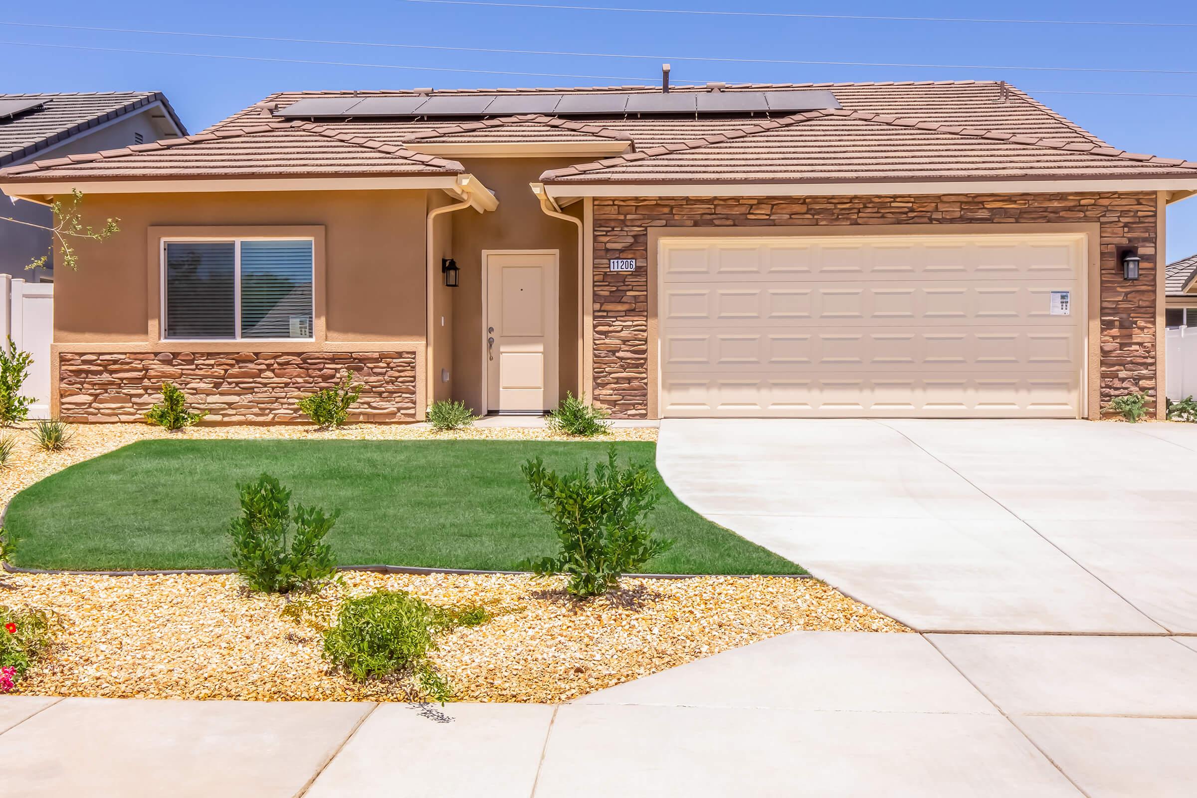 A modern single-story home with a stone facade and a tiled roof. The front yard features low-maintenance landscaping with grass and decorative gravel. A double garage is visible on the right, and a large window is positioned on the front of the house. The sky is clear and blue.