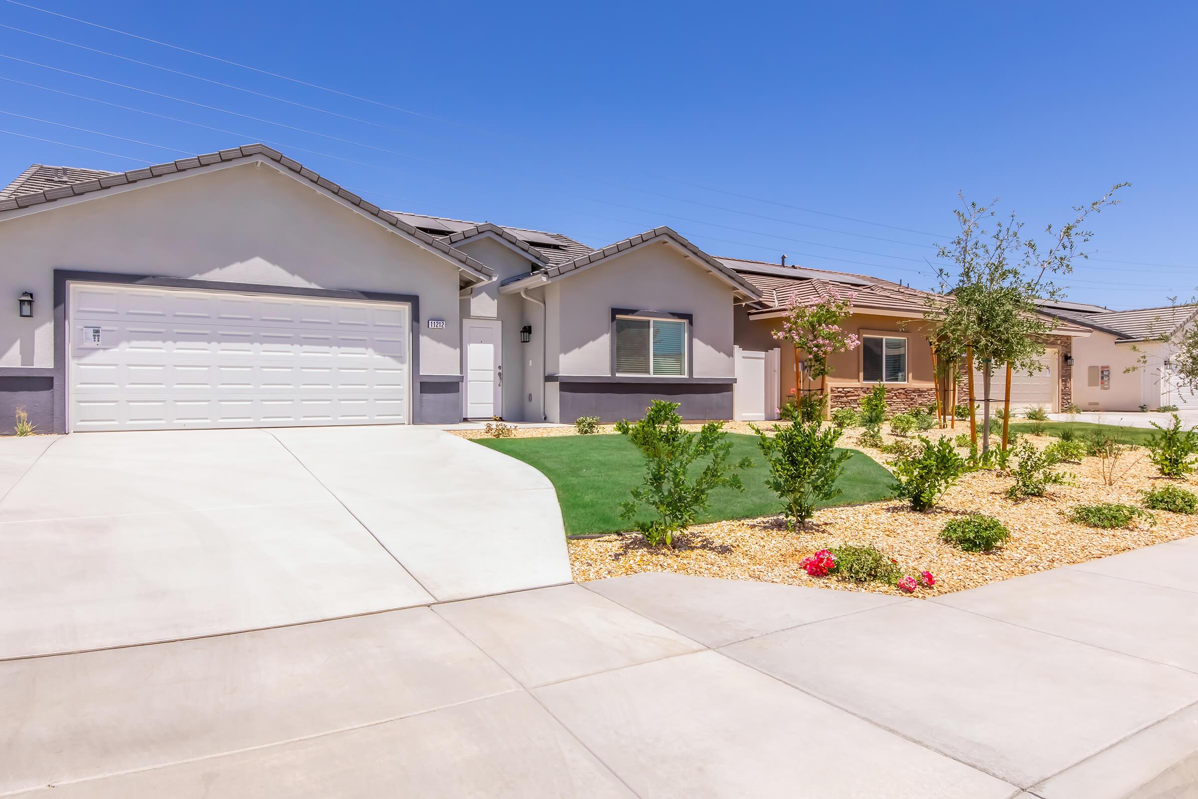 A row of modern residential houses with well-maintained exteriors and landscaped front yards. The driveways are paved, and there are small bushes and flowers in the garden areas. The sky is clear and blue, creating a bright and inviting atmosphere.