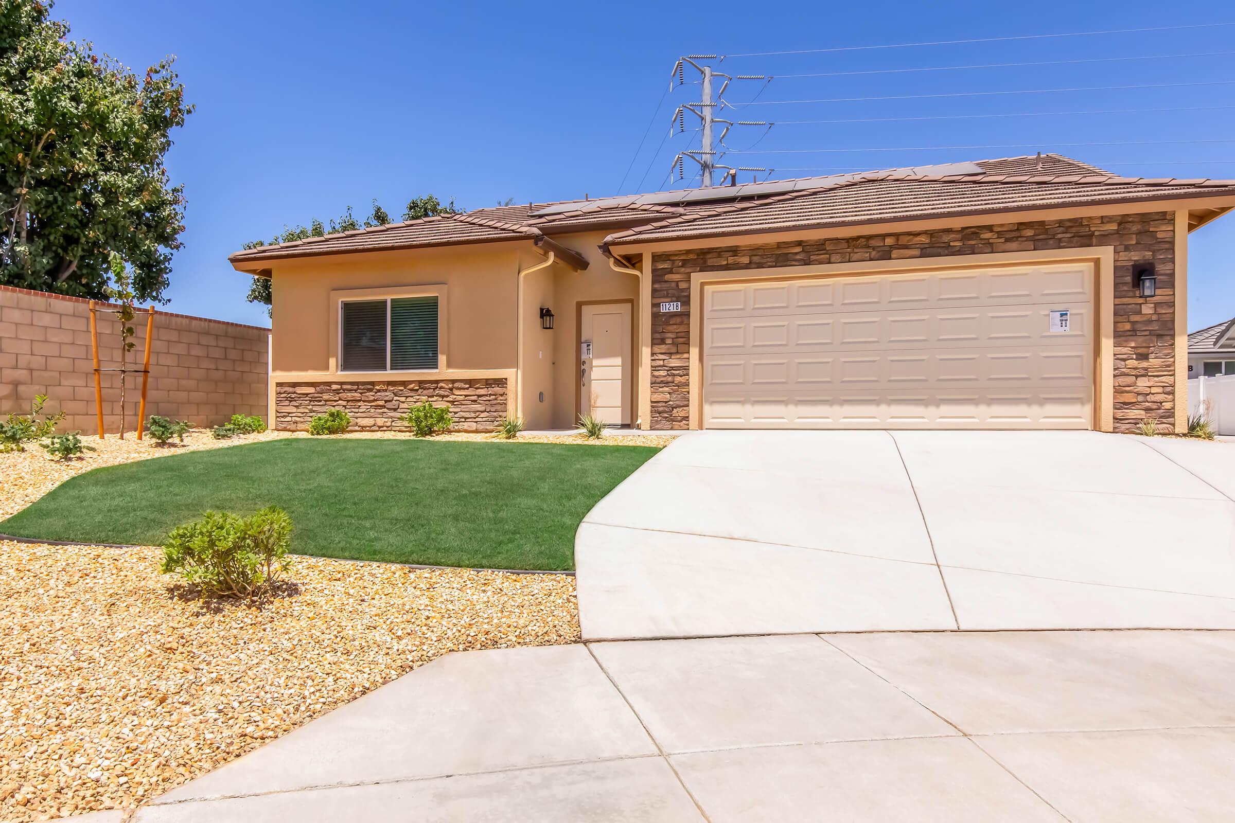 A single-story house with a stone-accented facade, a two-car garage, and well-maintained landscaping. The front yard features a low-maintenance design with gravel and small plants, set against a clear blue sky and utility poles in the background.