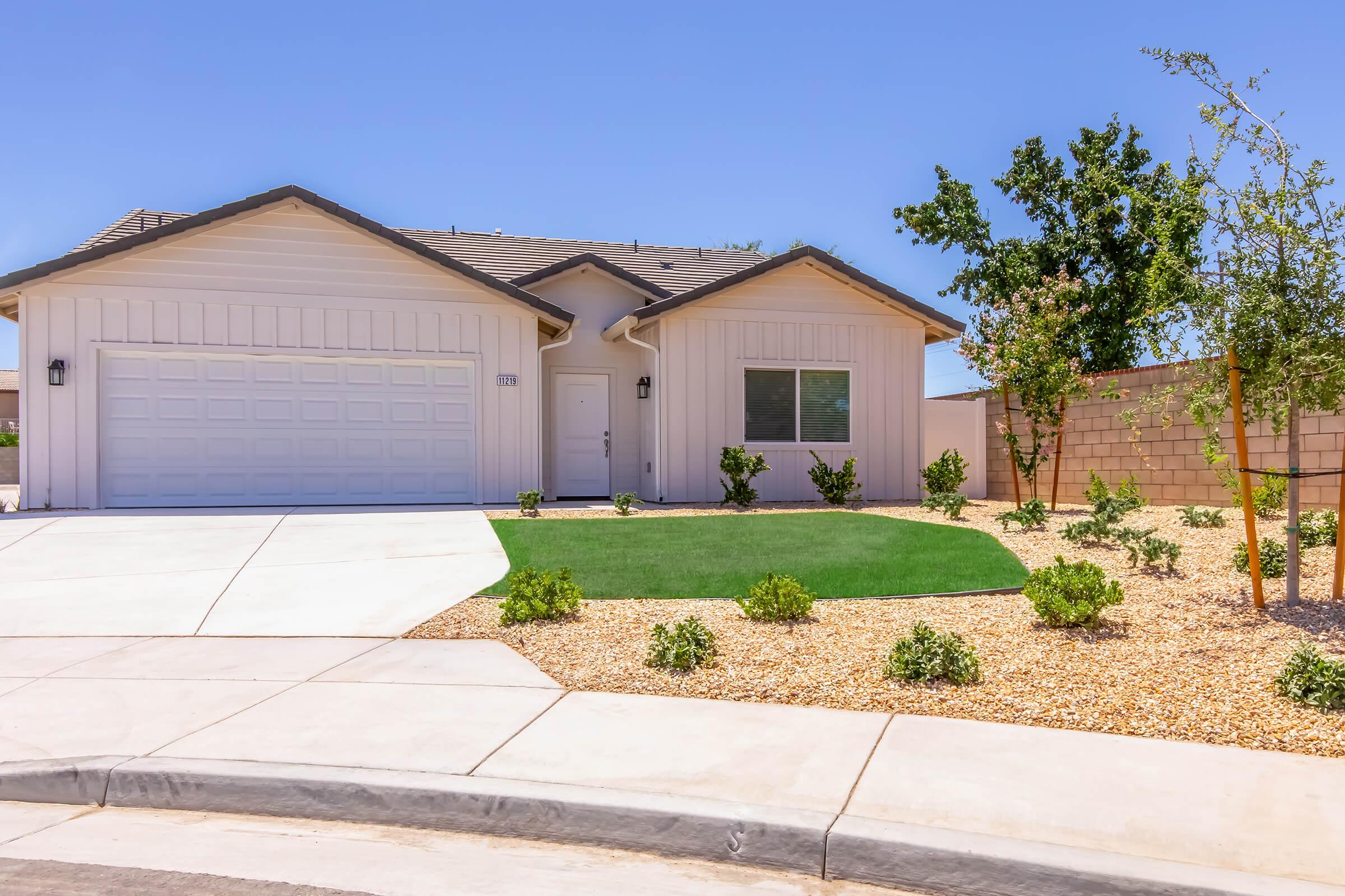 A modern single-story house with a light-colored exterior, a two-car garage, and a well-manicured front yard featuring small shrubs and decorative gravel. The house is set against a clear blue sky, with a few trees visible in the background, enhancing the appealing curbside view.
