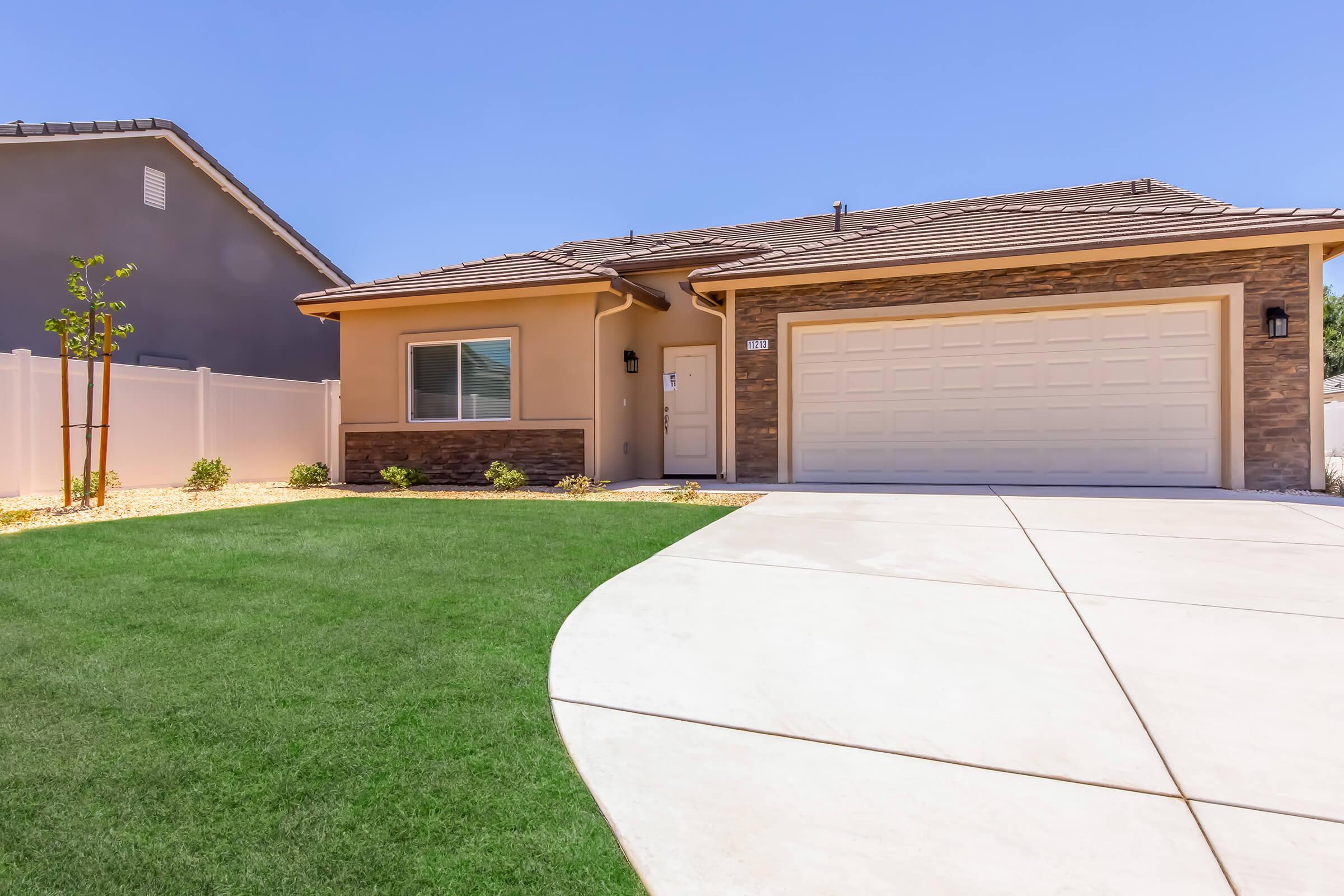 A modern single-story house with a tan exterior and stone accents. The front yard features well-maintained green grass, small shrubs, and a curved driveway leading to a two-car garage. A clear blue sky completes the scene, creating a bright and inviting atmosphere.