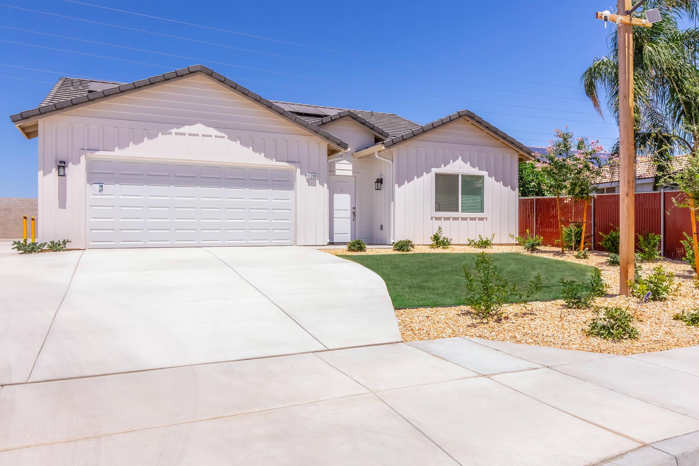 A modern single-story house with a white exterior, a two-car garage, and a neatly landscaped front yard featuring a small lawn and gravel. Clear blue skies and utility poles are visible in the background.
