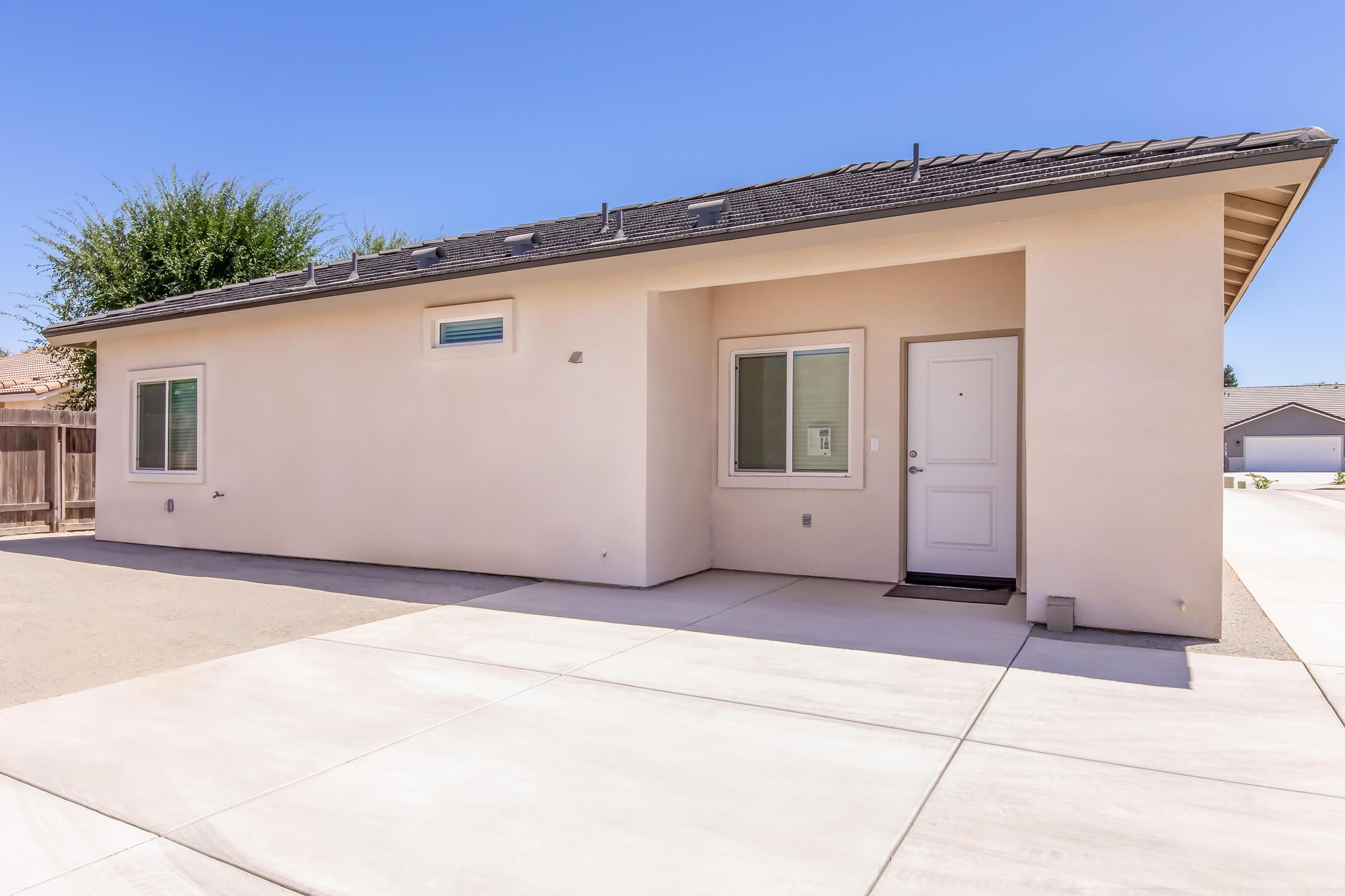 Single-story residential building with a light-colored exterior, large windows, and a front door. The surrounding area features a paved driveway and clear blue sky. The house has a simple, modern design, with minimal landscaping.