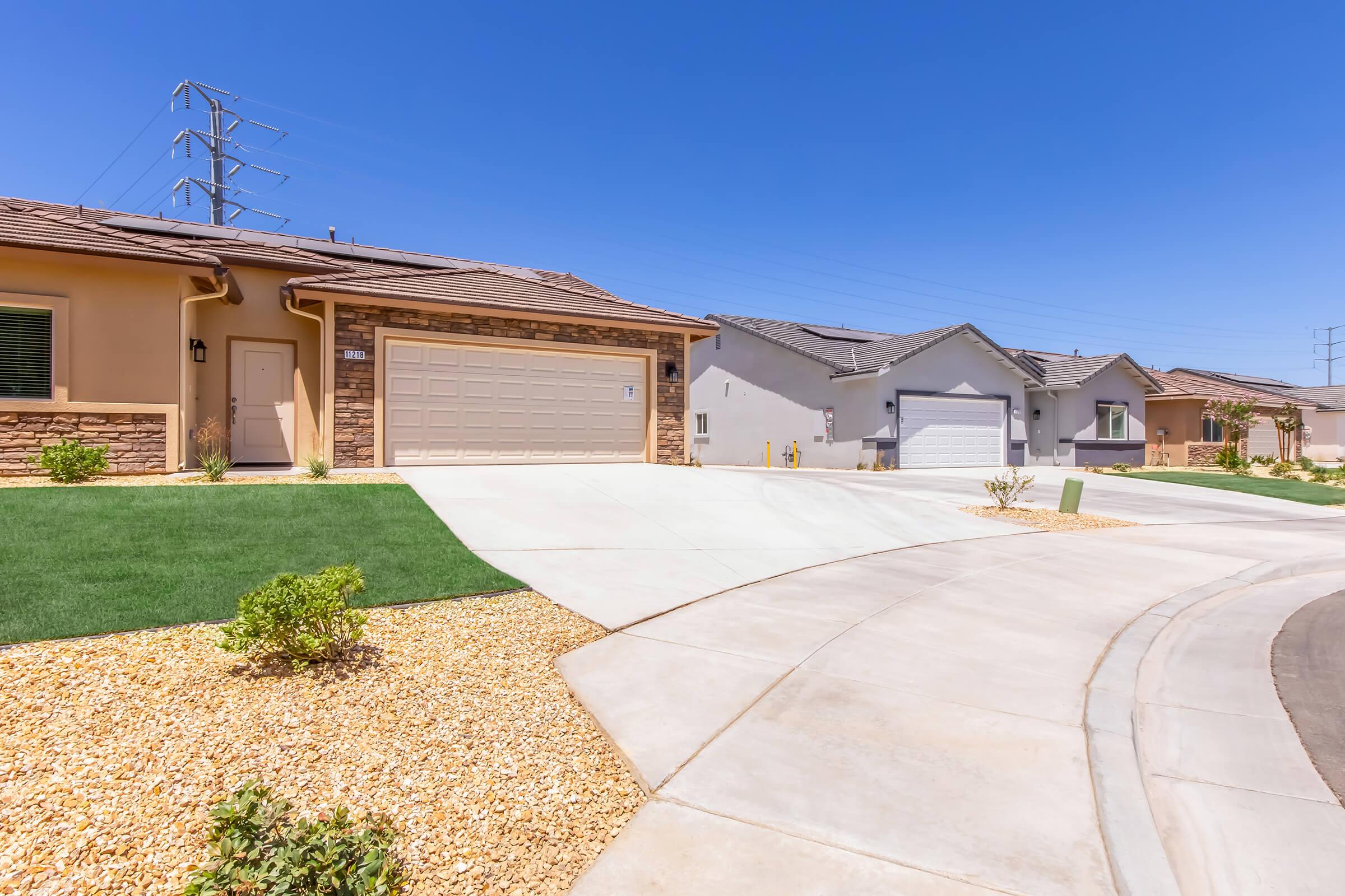A sunny residential street view featuring modern houses with light-colored exteriors, landscaped yards with small shrubs, and paved driveways. Power lines are visible in the background, and the sky is clear blue, creating a bright atmosphere.