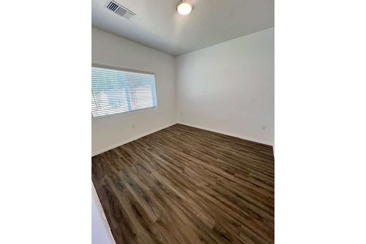 A spacious, empty room with light-colored walls and a wood-like floor. A window with blinds allows natural light to enter, while the ceiling is fitted with a simple light fixture. The overall ambiance is bright and airy, suitable for various uses.