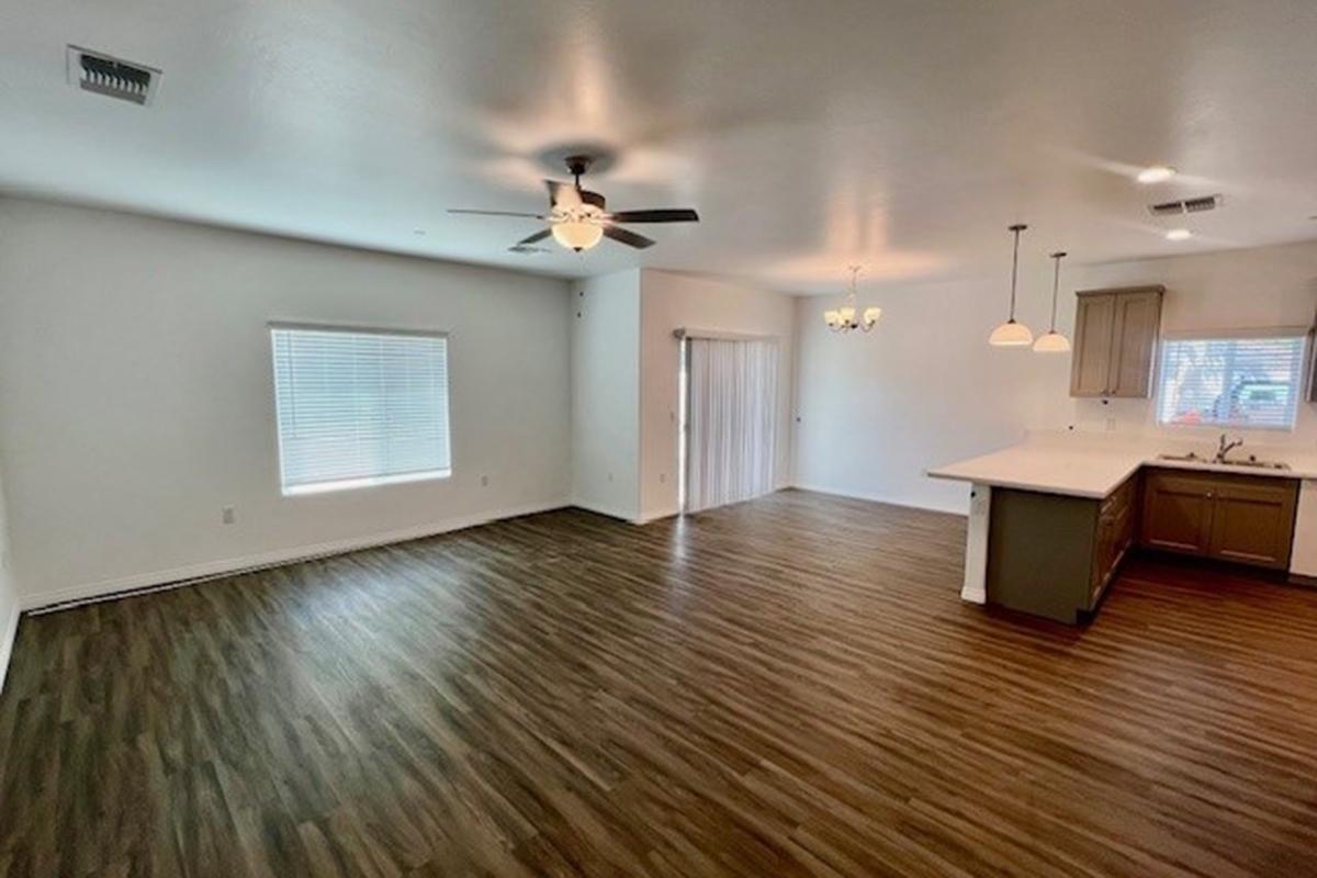 Spacious interior of a modern living area featuring wooden laminate flooring, a ceiling fan, and a large window with blinds. The open layout connects to a kitchen area, which includes a breakfast bar, and has natural light from overhead fixtures. A sliding door leads to an outdoor space.
