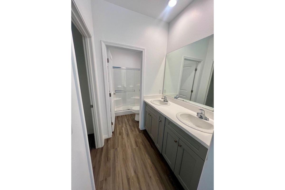 A modern bathroom featuring two sinks with mirrors above, gray cabinetry, and a neutral color scheme. The space includes a frosted glass shower enclosure and light wood-like flooring, providing a clean and spacious appearance.