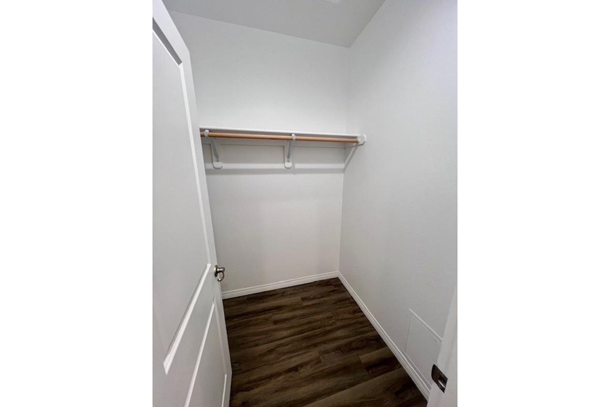 A clean, empty closet featuring white walls and a wooden floor. The closet has a single shelf mounted against the back wall and is illuminated by natural light. The door is partially open, and there are no items inside, creating a minimalist and spacious appearance.