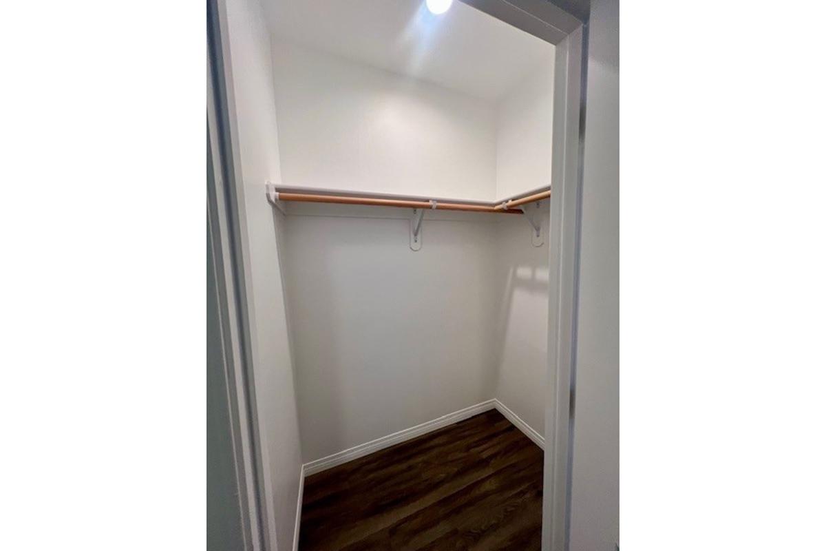 A clean, empty closet space with two wooden shelves mounted on the wall. The walls are painted white, and there is warm wooden flooring. A ceiling light illuminates the area, creating a bright and inviting atmosphere.