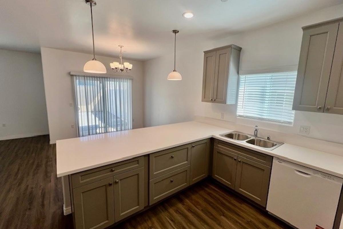 A modern kitchen with gray cabinetry, a white countertop, and stainless steel sink. There are two pendant lights above the counter, and a large window with blinds providing natural light. A dining area with a light fixture is in the background, and the flooring is dark wood.