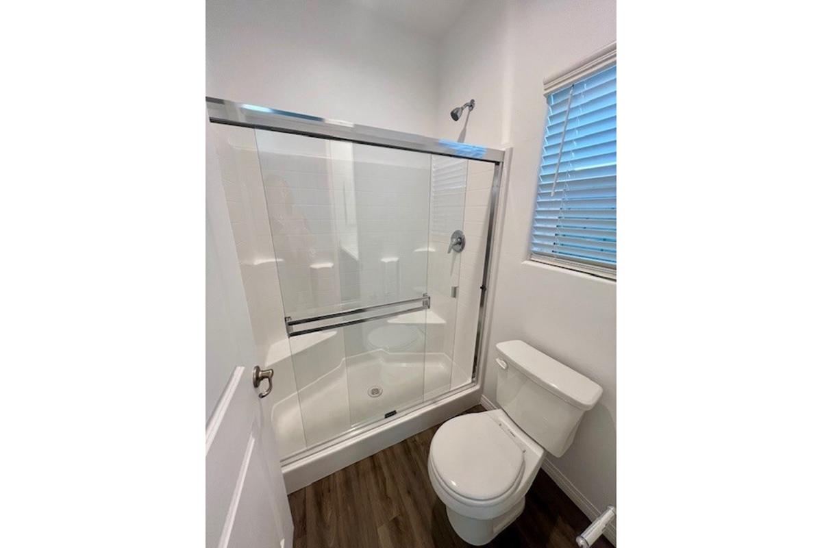 A modern bathroom featuring a glass shower enclosure, a white toilet, and a window with blinds. The walls are painted white, and the flooring is dark wood. The overall design is clean and minimalist.