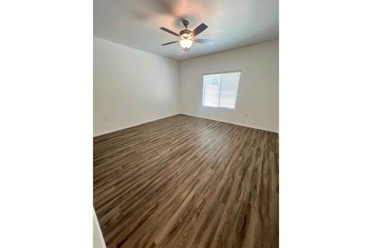 A spacious, empty room featuring light-colored walls, a ceiling fan, and a window with blinds. The floor is covered in wood-like laminate, creating a warm and inviting atmosphere. Ideal for various uses such as a living area or bedroom.