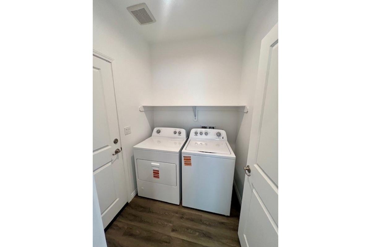 A compact laundry room featuring a stacked washer and dryer set on the left and a dryer on the right. The walls are painted white, with a shelf above the appliances for storage. There is a door on the left side leading to another room, and the flooring has a wood-like appearance.