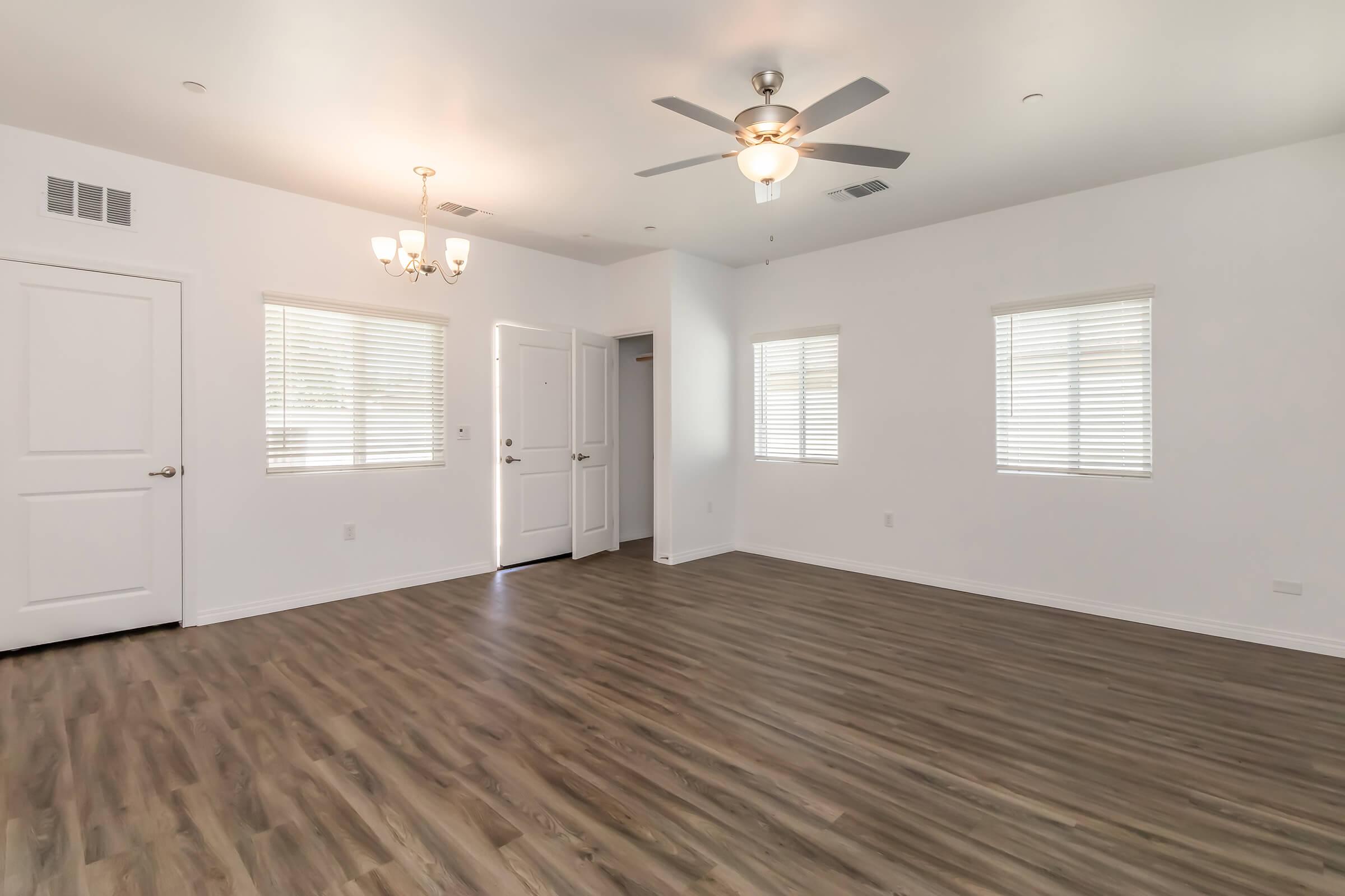 A spacious, well-lit room with light-colored walls and a wooden floor. It features two windows with blinds, a door leading outside, and a ceiling fan. A chandelier hangs in the center, adding elegance to the space. The overall atmosphere is bright and open, suitable for various furniture arrangements.