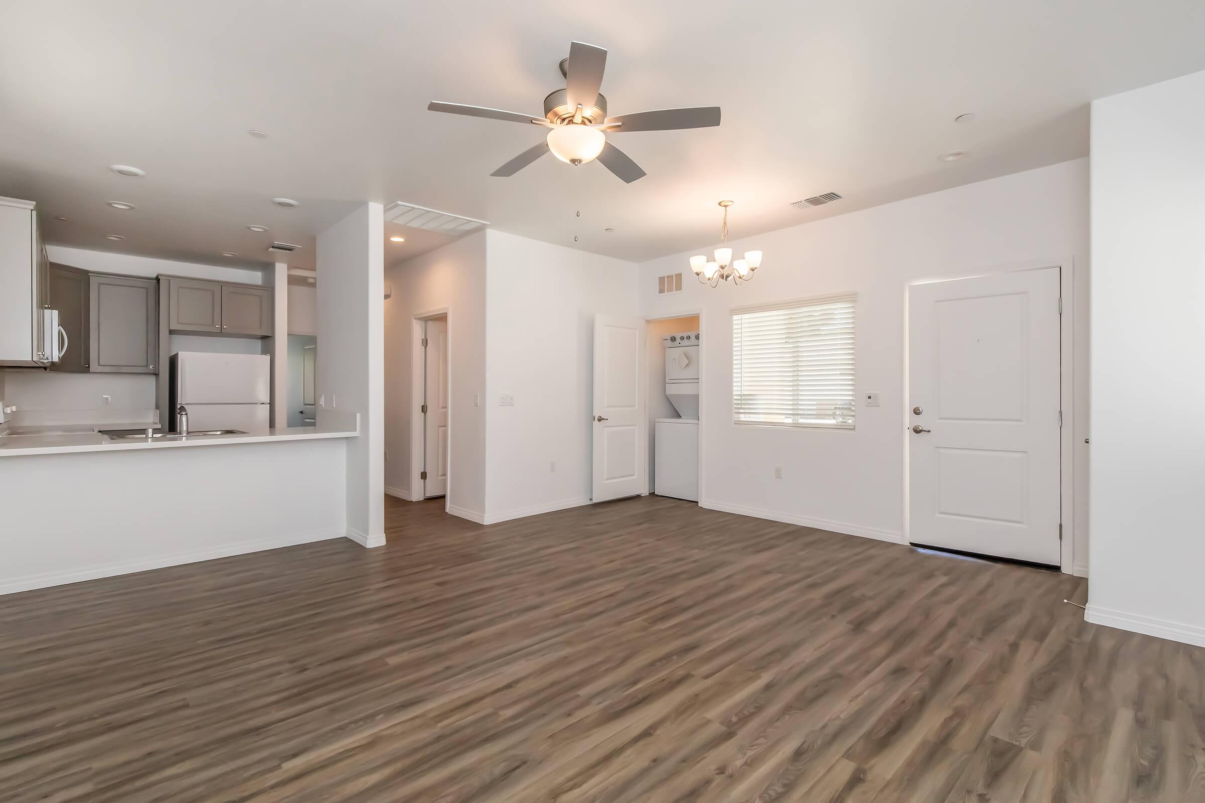 Spacious and modern living area with laminate flooring, ceiling fan, and natural light from windows. The open layout includes a kitchen area with cabinetry and appliances, a dining space, and a front door leading outside. A laundry area is visible in the background.