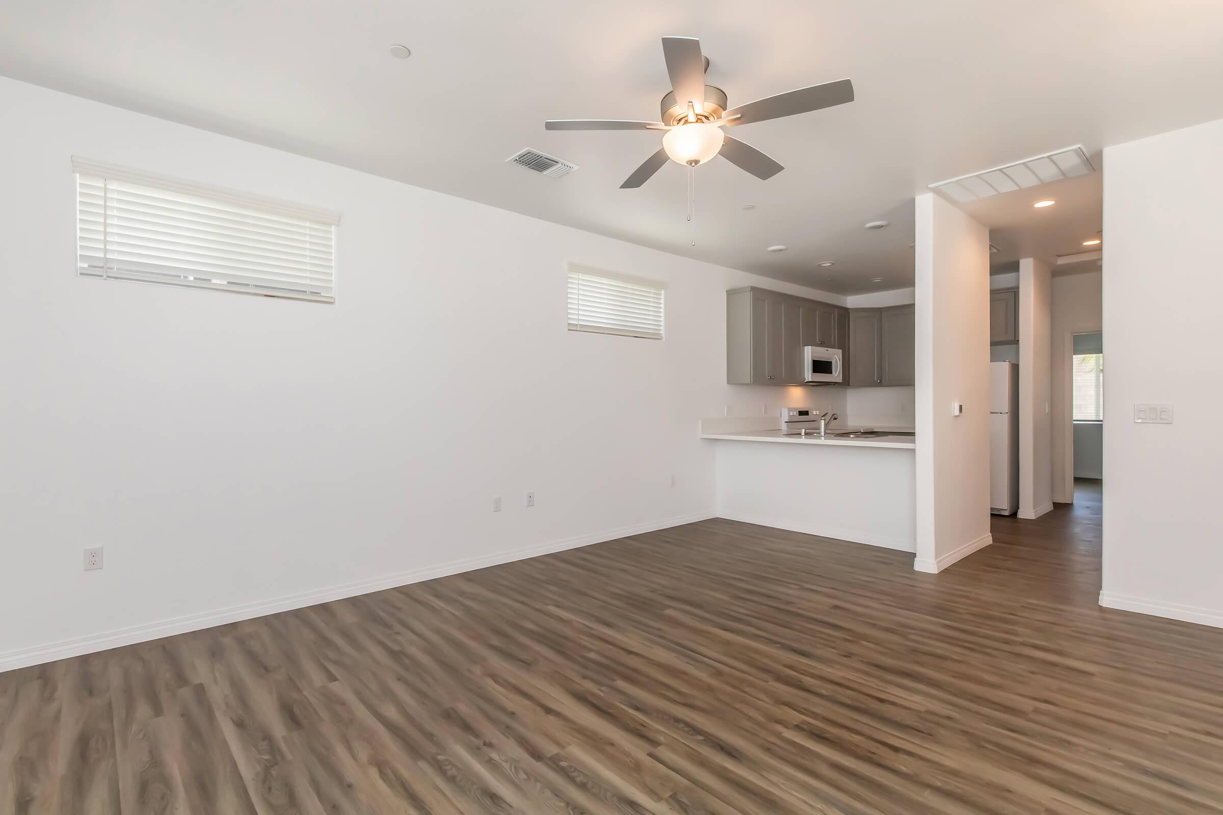 Bright, modern living area with light wood-look floors, ceiling fan, and two small windows letting in natural light. A kitchen with gray cabinets and white countertops is partially visible in the background, alongside a doorway leading to another room. The walls are painted white, creating a clean and open feel.