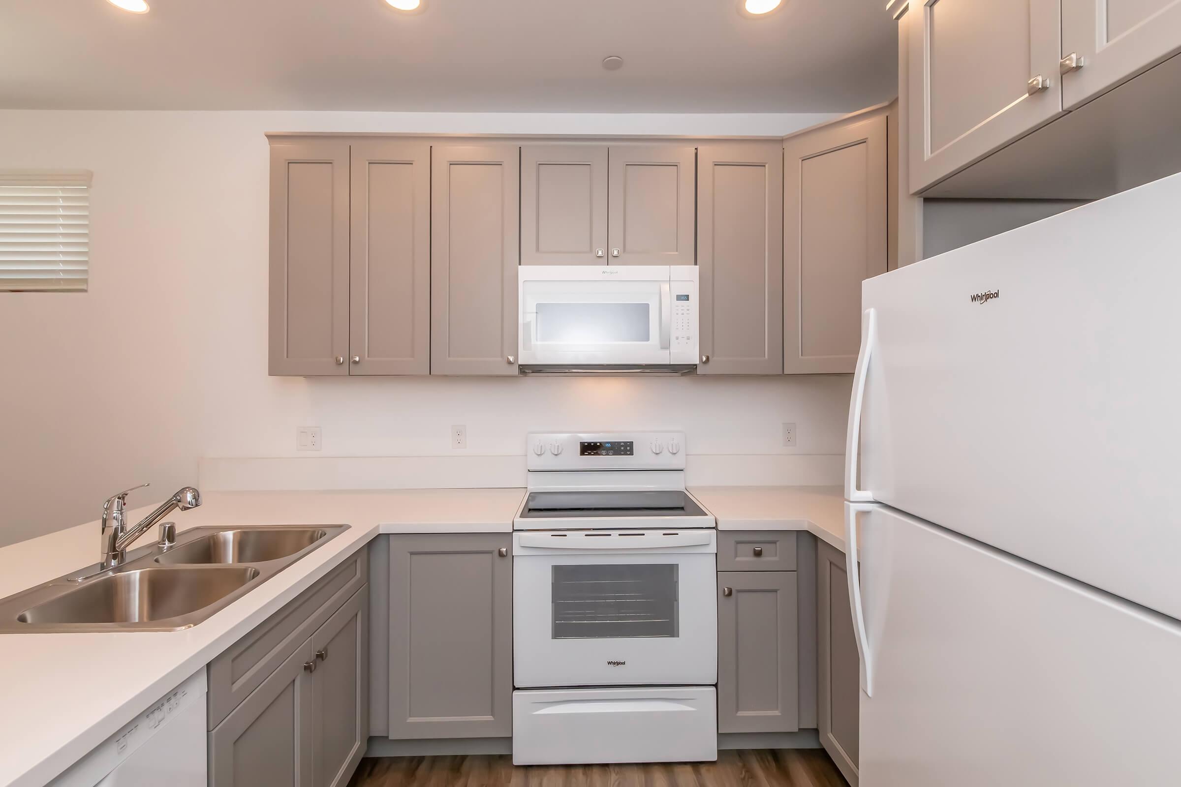 Modern kitchen featuring light gray cabinetry, a white microwave and stove, and a white refrigerator. The countertops are light-colored, and there are dual sinks. The room is well-lit with overhead lights and has a clean, contemporary design.