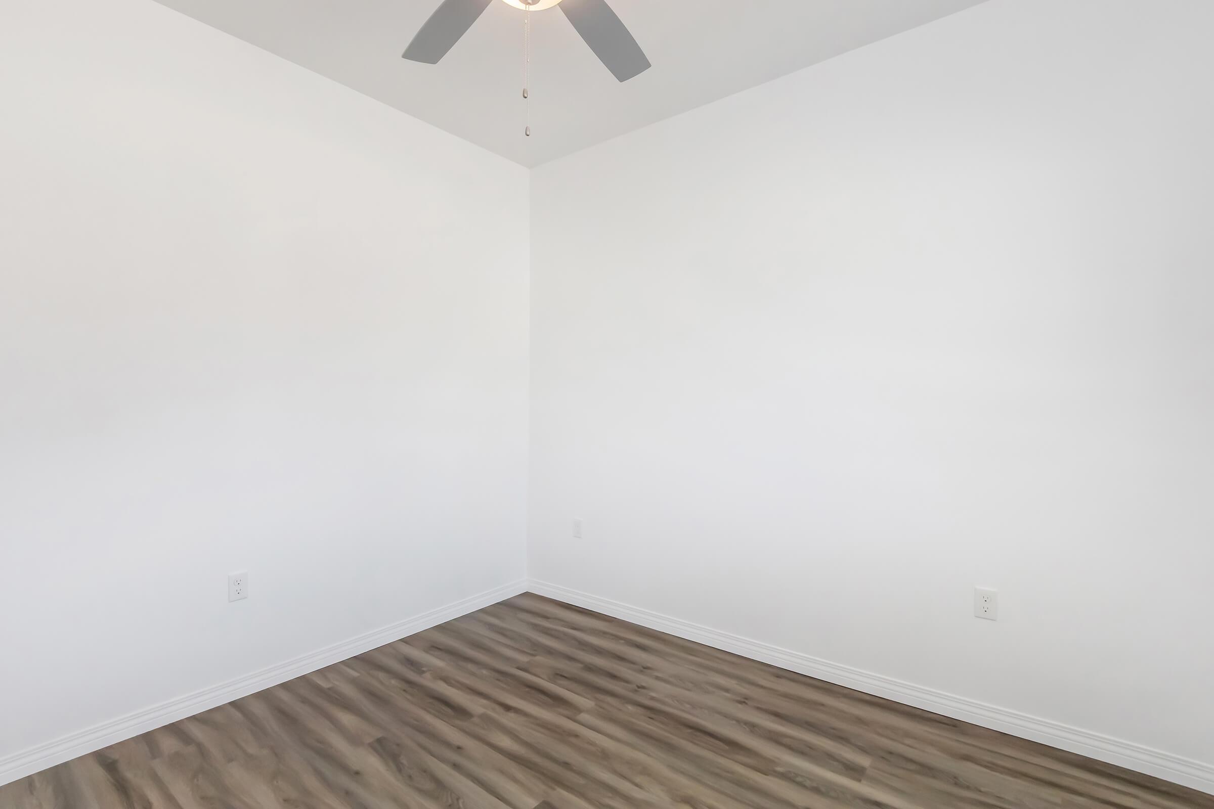 Empty room with white walls and a ceiling fan. The flooring is light brown with a wood-like finish. There are no furnishings or decorations, creating a minimalist appearance. The room features two electrical outlets on the wall. Natural light may enter through an unseen window.