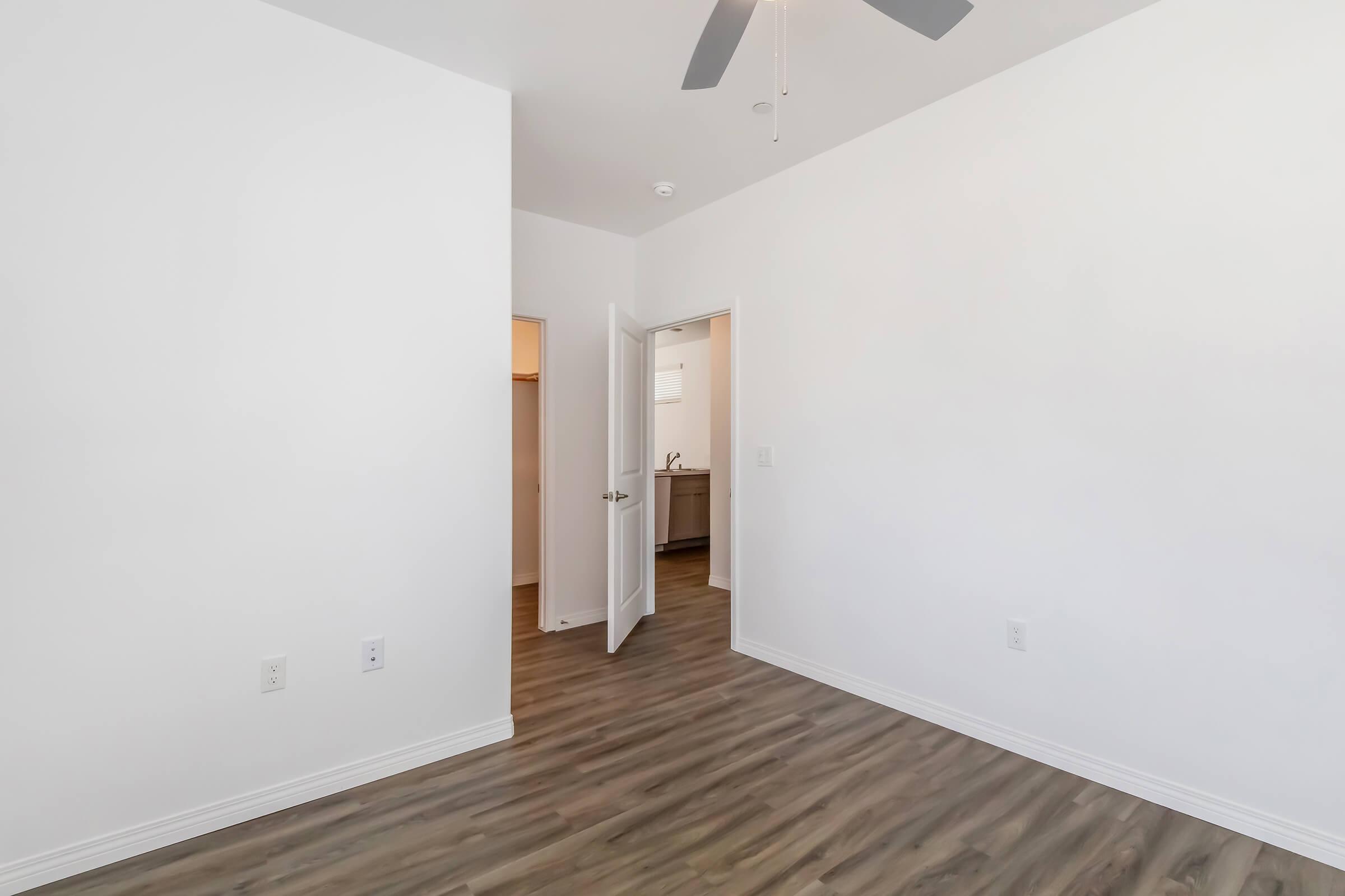 A bright, empty room with white walls and a ceiling fan. The floor is covered with light-colored wooden planks. There are two doorways leading to adjacent spaces, one with a door partially open, suggesting easy access to the connected rooms. Overall, the space feels airy and modern.