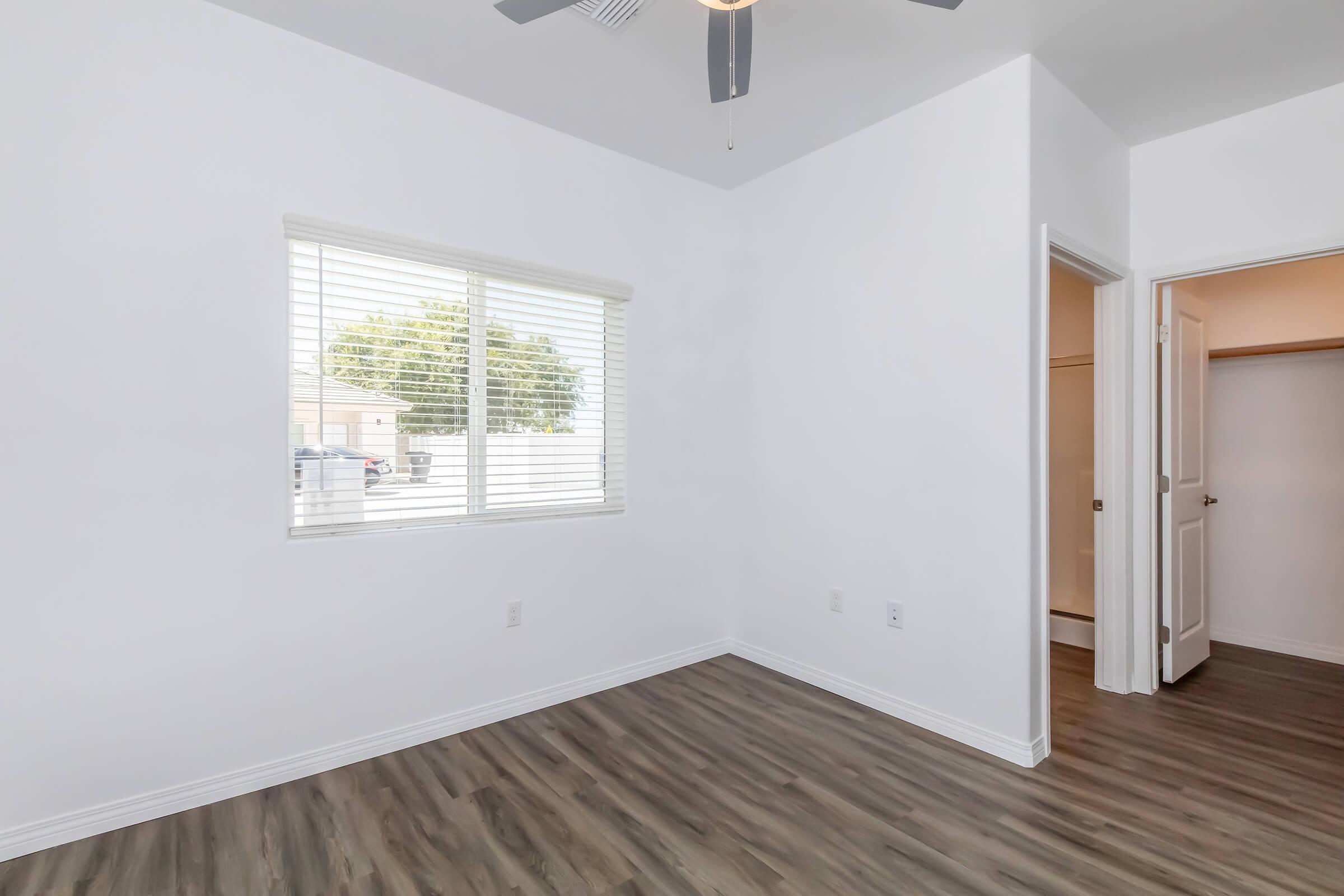 A bright, empty room with white walls and a ceiling fan. A window with blinds allows natural light to enter, and a door leads to a closet. The floor features wood-like laminate, giving a modern touch to the space. The overall atmosphere is clean and airy, ideal for personal customization.