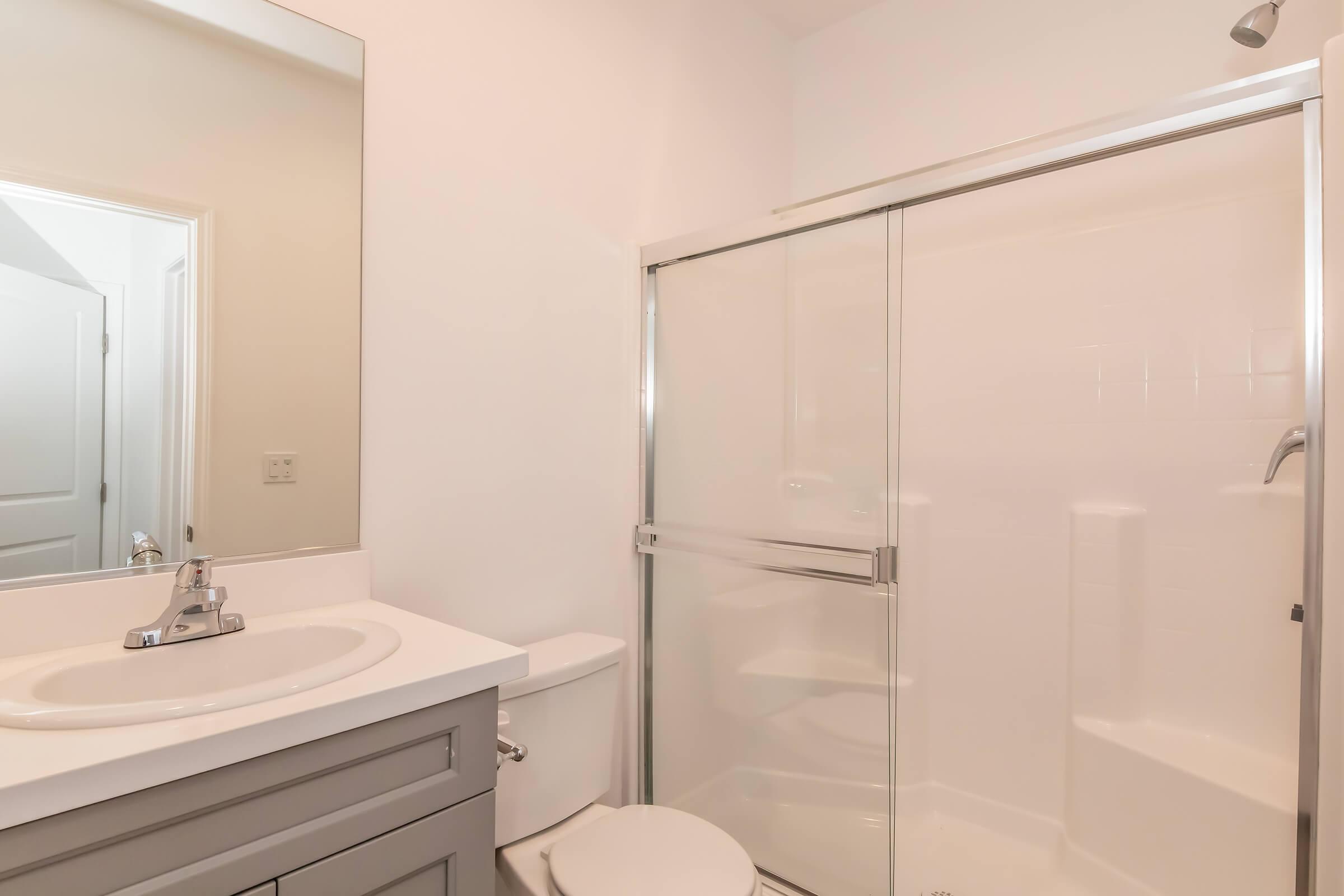 A modern bathroom featuring a white shower enclosure with glass doors, a small vanity with a sink, a toilet, and a large mirror. The walls are painted in a light color, creating a bright and clean atmosphere. The space appears well-maintained and contemporary.