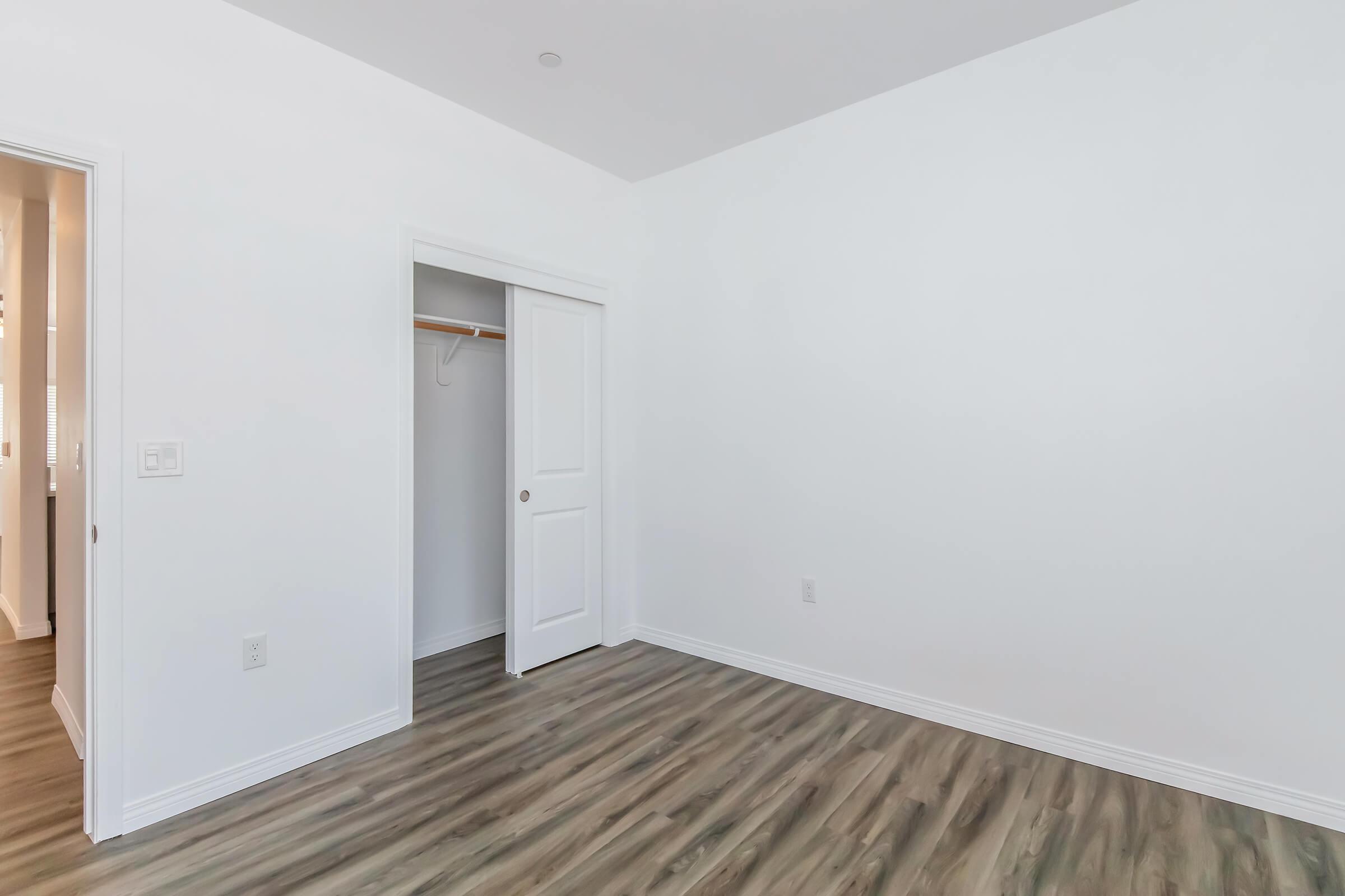 Empty room with white walls and a light-colored wooden floor. There is a closet with a door on the left side. The room is well-lit with natural light, and the overall layout is simple and minimalist, making it suitable for various design choices.
