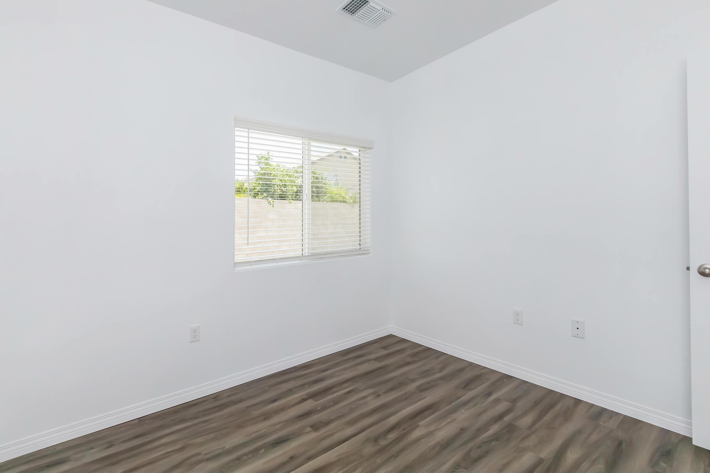 A minimalist empty room with white walls and a window featuring horizontal blinds. The floor is a light brown wooden design, and there are unobtrusive electrical outlets and switches on the walls. Natural light enters through the window, highlighting the spaciousness of the room.