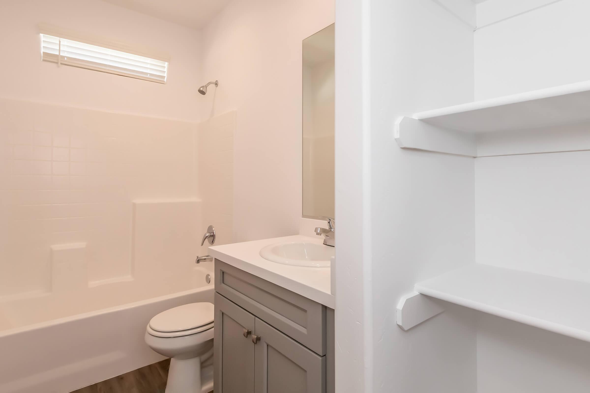 A clean and modern bathroom featuring a white bathtub, a sink with a gray cabinet, a mirror, and a small window. There is also an open shelf for storage, and the flooring appears to be a wood-like material, giving the space a contemporary feel.
