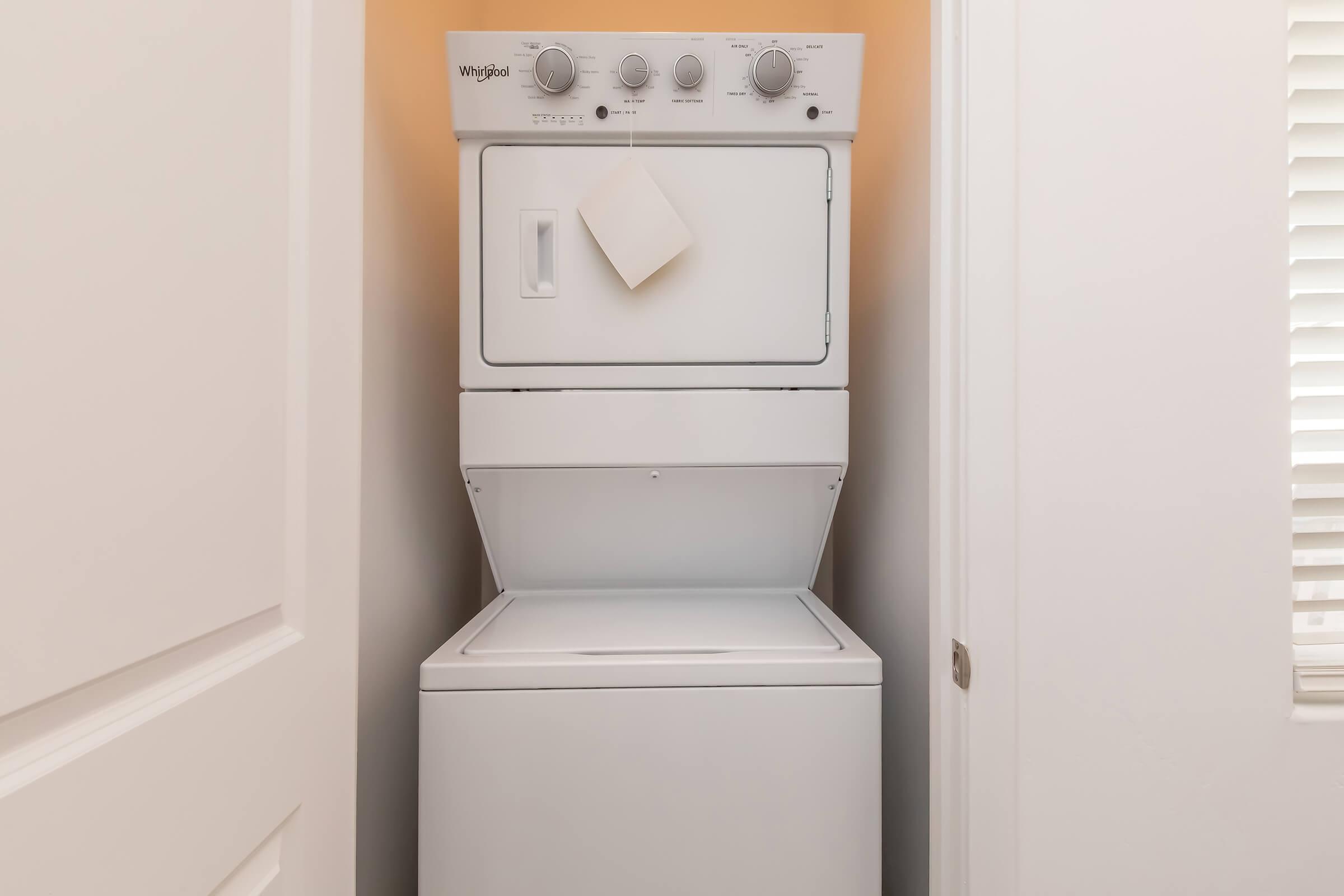 A stacked washer and dryer unit in a small laundry closet. The machines are white, with control knobs on the top and a blank piece of paper attached to the dryer. The closet has light-colored walls and a door partially visible on the right, along with a window providing natural light.