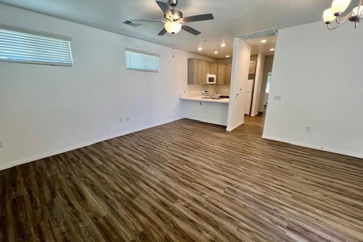 A spacious, light-filled living area with wooden flooring, showcasing a ceiling fan and two windows with blinds. A doorway leads to a modern kitchen with light-colored cabinetry in the background. The walls are painted white, creating a bright and airy atmosphere.
