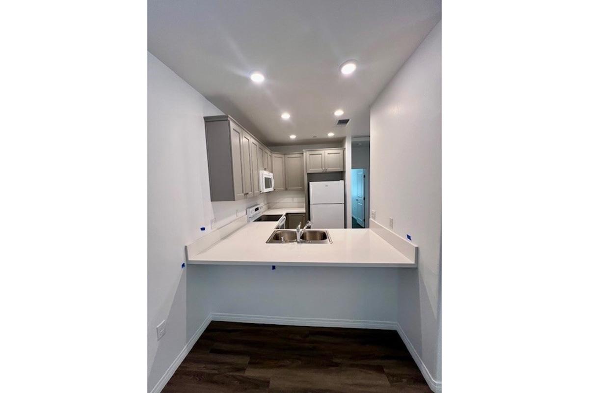 A modern kitchen featuring light gray cabinets, a white countertop, and an open layout. The kitchen has stainless steel appliances, including a microwave and a refrigerator, and is illuminated by recessed lighting. The flooring is a dark wood finish, providing contrast to the bright walls.