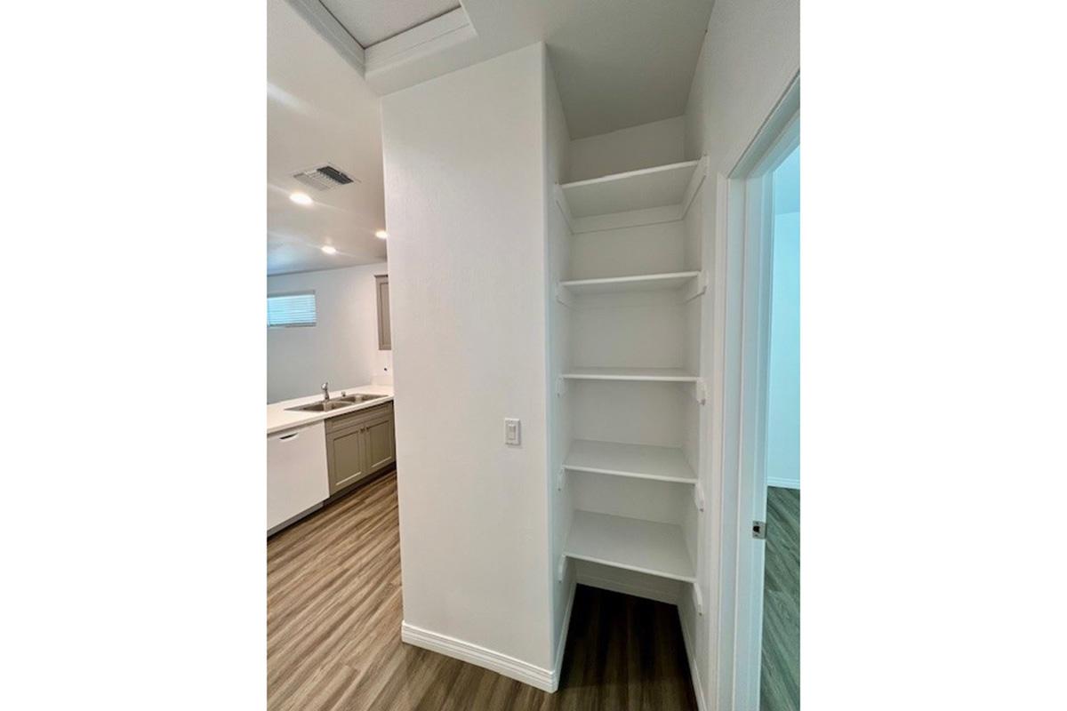 A corner of a modern kitchen featuring a white wall with open shelves for storage. The space is well-lit with recessed lighting, and there are wood-like floors. The kitchen area is visible in the background, showcasing cabinets and a sink.