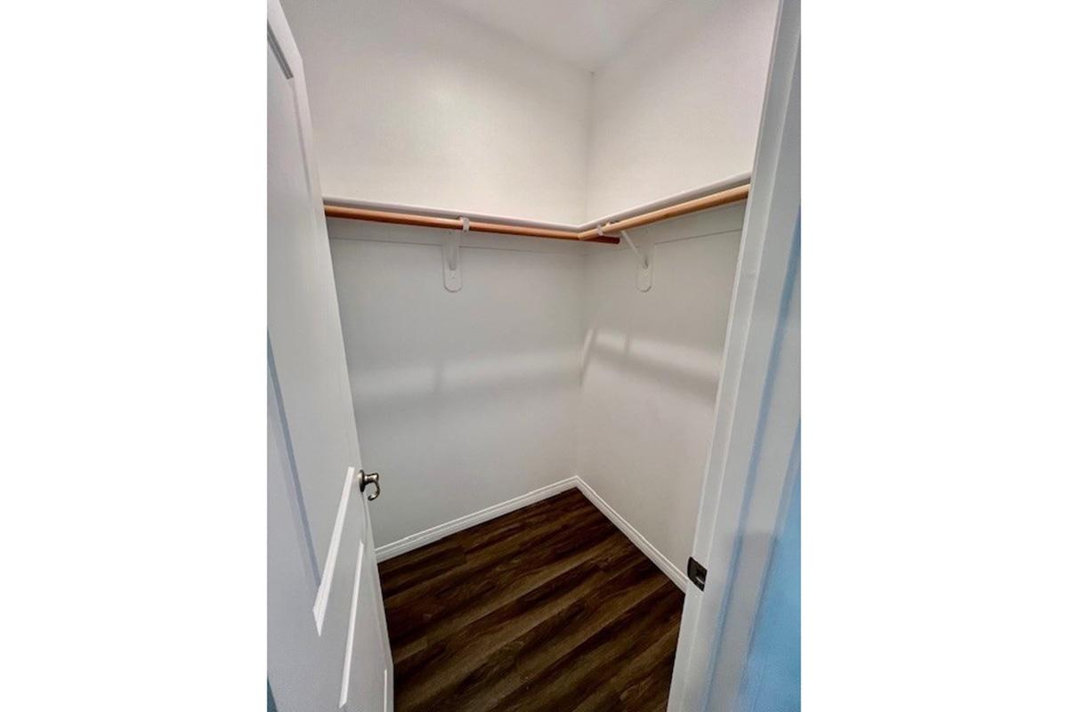 Empty closet featuring a wooden floor, white walls, and a single shelf with hanging rod. The closet is well-lit and has two doors, one partially open, giving a clear view of the interior space.