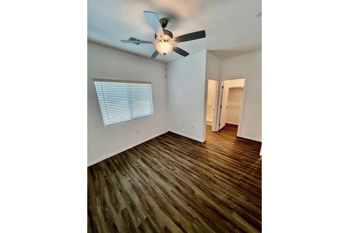 A bright, spacious room featuring wooden flooring, a ceiling fan with light, and a window with blinds. A doorway leads to a closet outside of the frame, enhancing the room's natural light and open feel. The walls are painted in a light color, adding to the airy atmosphere.