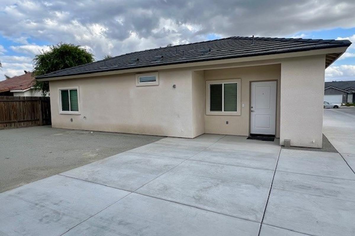 A single-story residential house with a light-colored exterior, featuring two windows on the front and a white door. The driveway is made of concrete, with a clear view of the surrounding area under a partly cloudy sky. The yard is gravel with no landscaping visible.