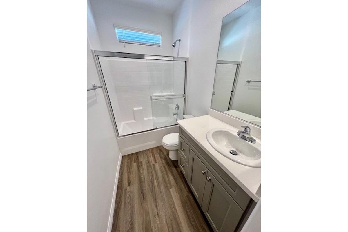 A modern bathroom featuring a glass shower enclosure, a white bathtub, a clean toilet, and a single sink vanity with storage. The walls are painted in white, and the floor has light wood-like tiles. A small window allows natural light into the space.