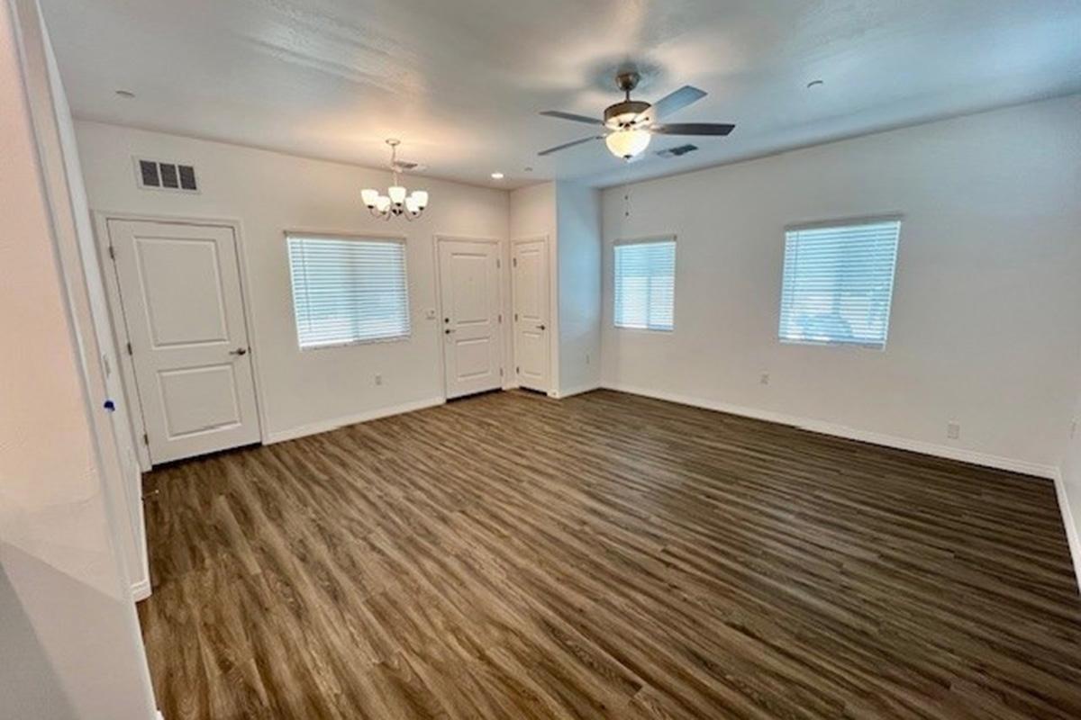 A spacious, well-lit room with a ceiling fan and light fixtures. The walls are painted white, and there are three windows providing natural light. The flooring is a wooden texture. There are two doors visible, suggesting access to other areas. The overall ambiance is clean and contemporary.