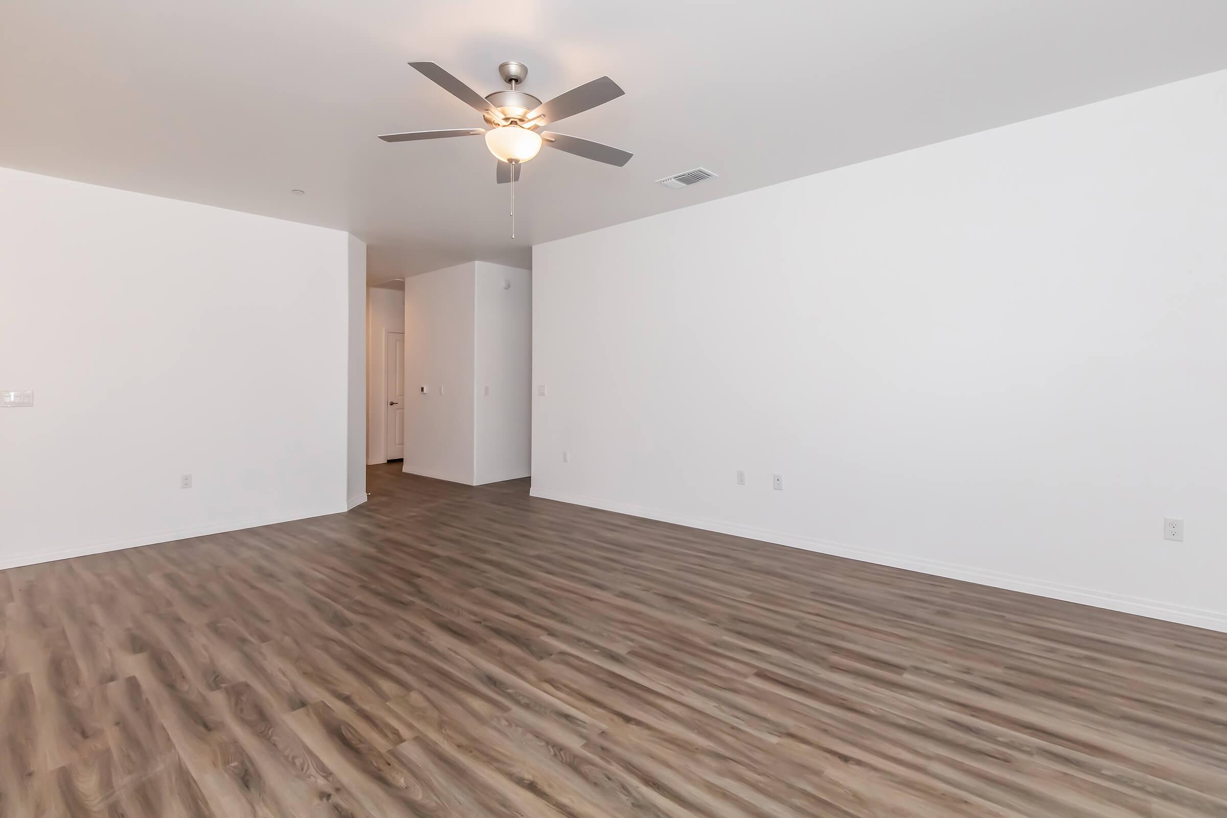 Spacious interior of a modern room featuring light-colored walls, a ceiling fan, and wood-style flooring. The space is empty, giving a sense of openness, with a hallway visible on the right leading to other rooms. Ideal for planning furniture layout or design.