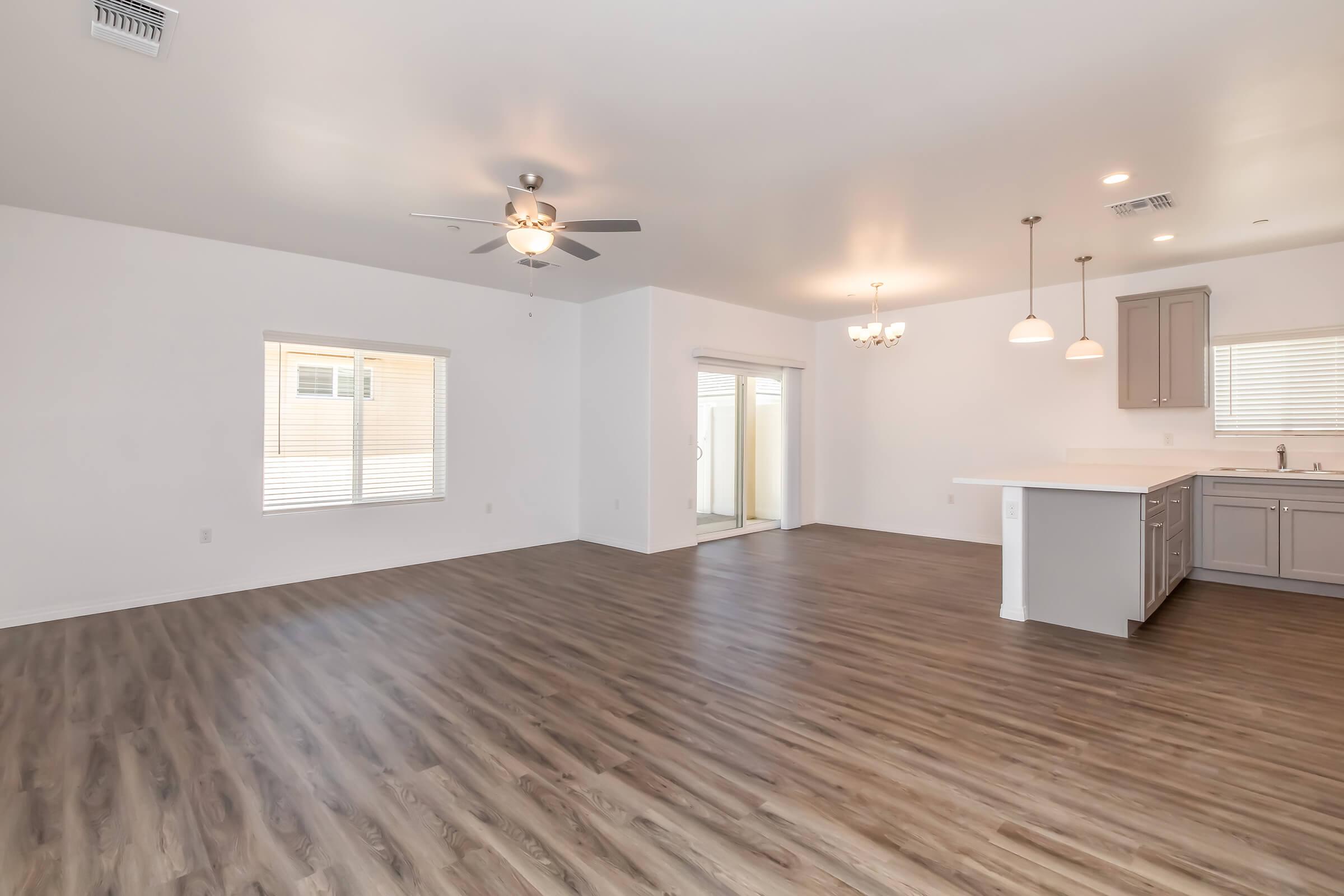 A spacious, modern living area featuring light-colored walls and wood-style flooring. On one side, there’s a large window letting in natural light, while on the other, a kitchen with light cabinetry is visible. The room is well-lit with ceiling lights and a ceiling fan, creating a warm, inviting atmosphere.
