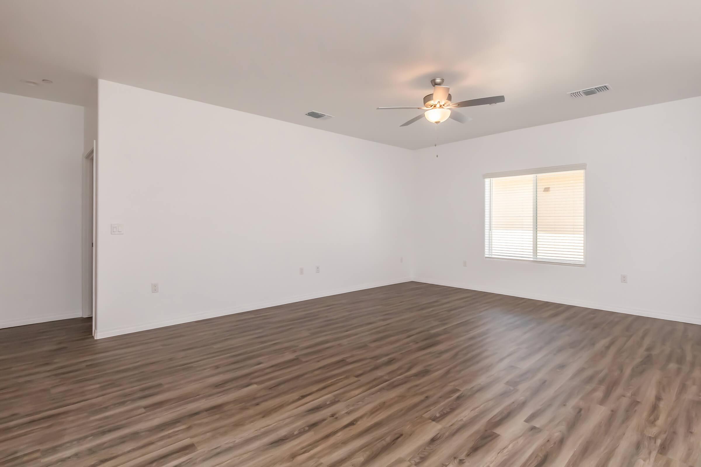 Spacious interior of a room with light-colored walls and hardwood flooring. A ceiling fan is installed, and there is a large window allowing natural light to enter. The room appears empty and is designed for versatility in layout and decor.