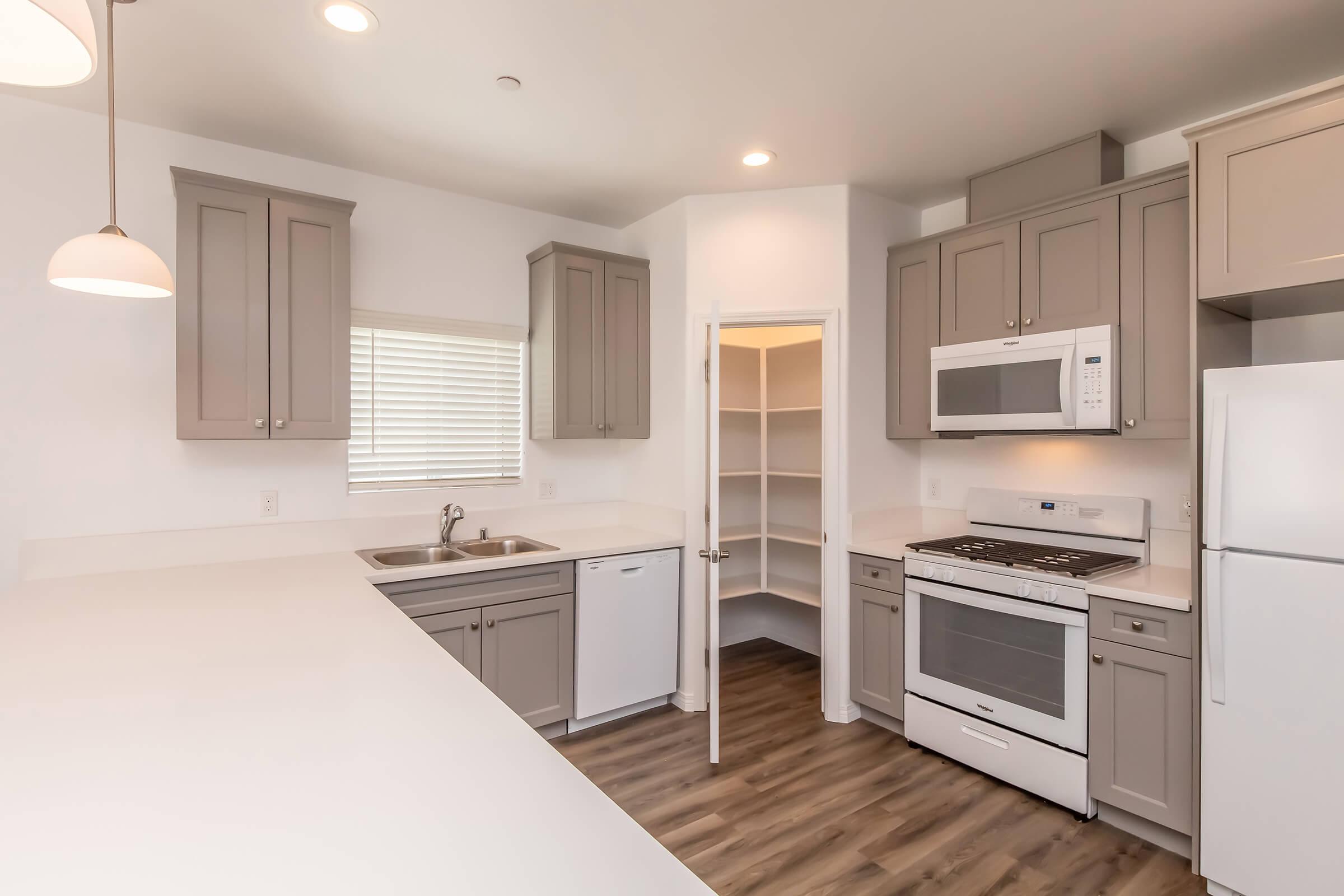 A modern kitchen featuring light gray cabinets, a countertop with a sink and dishwasher, a gas stove with an oven, and a microwave. A pantry area is visible through an open door, along with natural light coming in from a window. The floor has a wood-like finish.