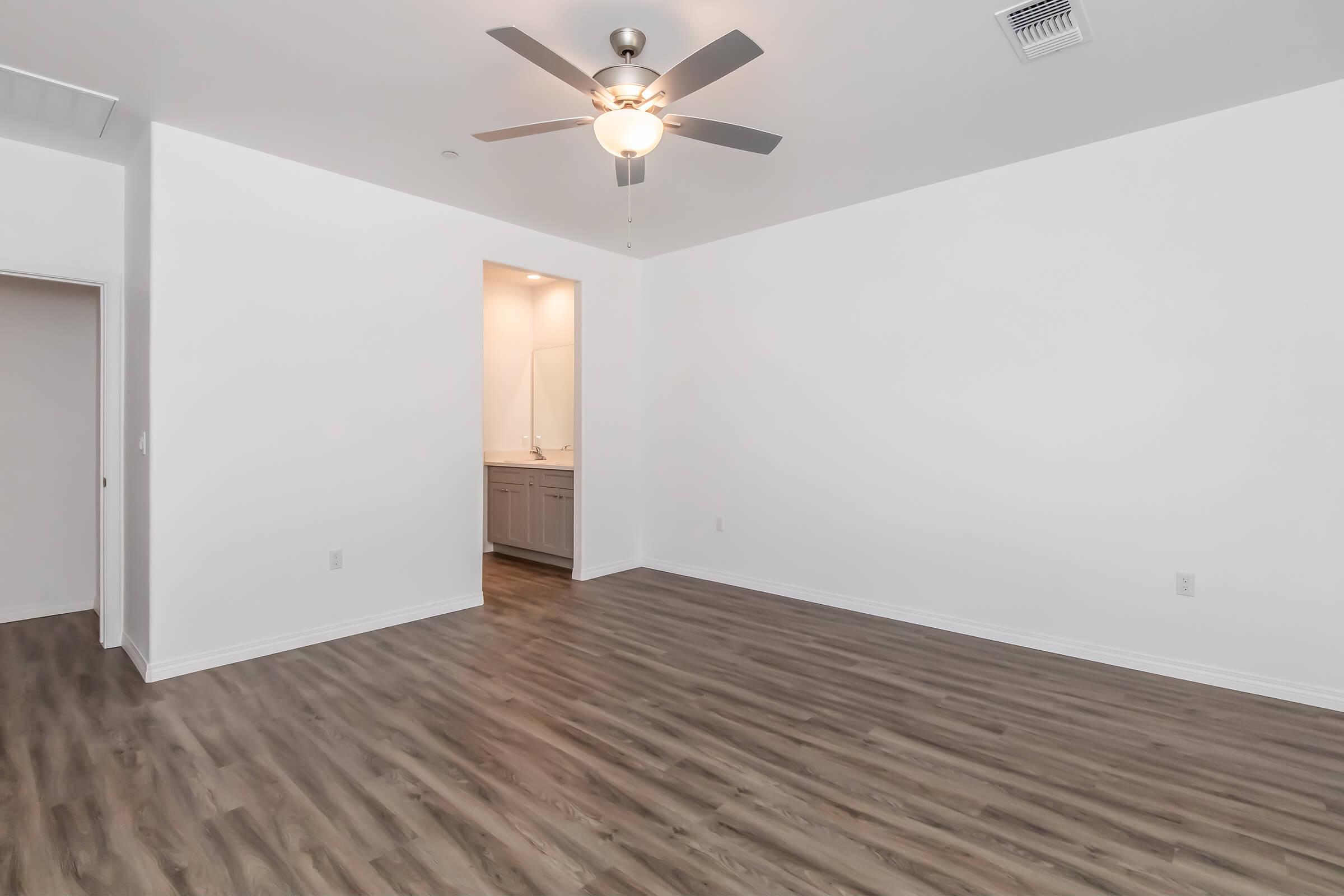 A spacious, empty room with light wood flooring, a ceiling fan, and white walls. There is an opening leading to a bathroom area with a vanity visible in the background. The room is well-lit and has a modern, clean aesthetic.