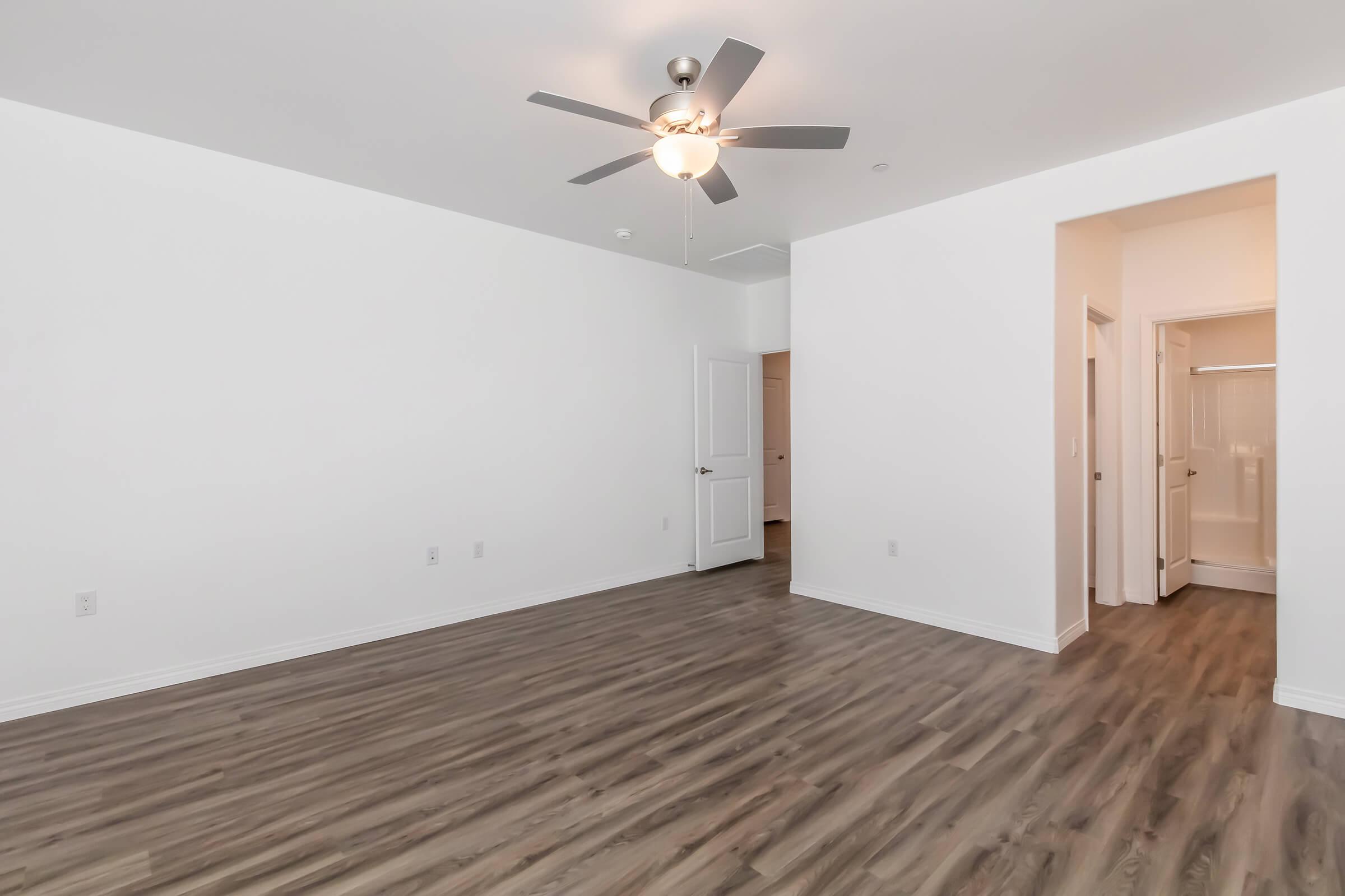 Spacious, well-lit room with light wood flooring and white walls. A ceiling fan is mounted overhead. There is a closed door leading to another space, and to the right, an open doorway reveals a bathroom. The room appears empty and ready for furnishing.